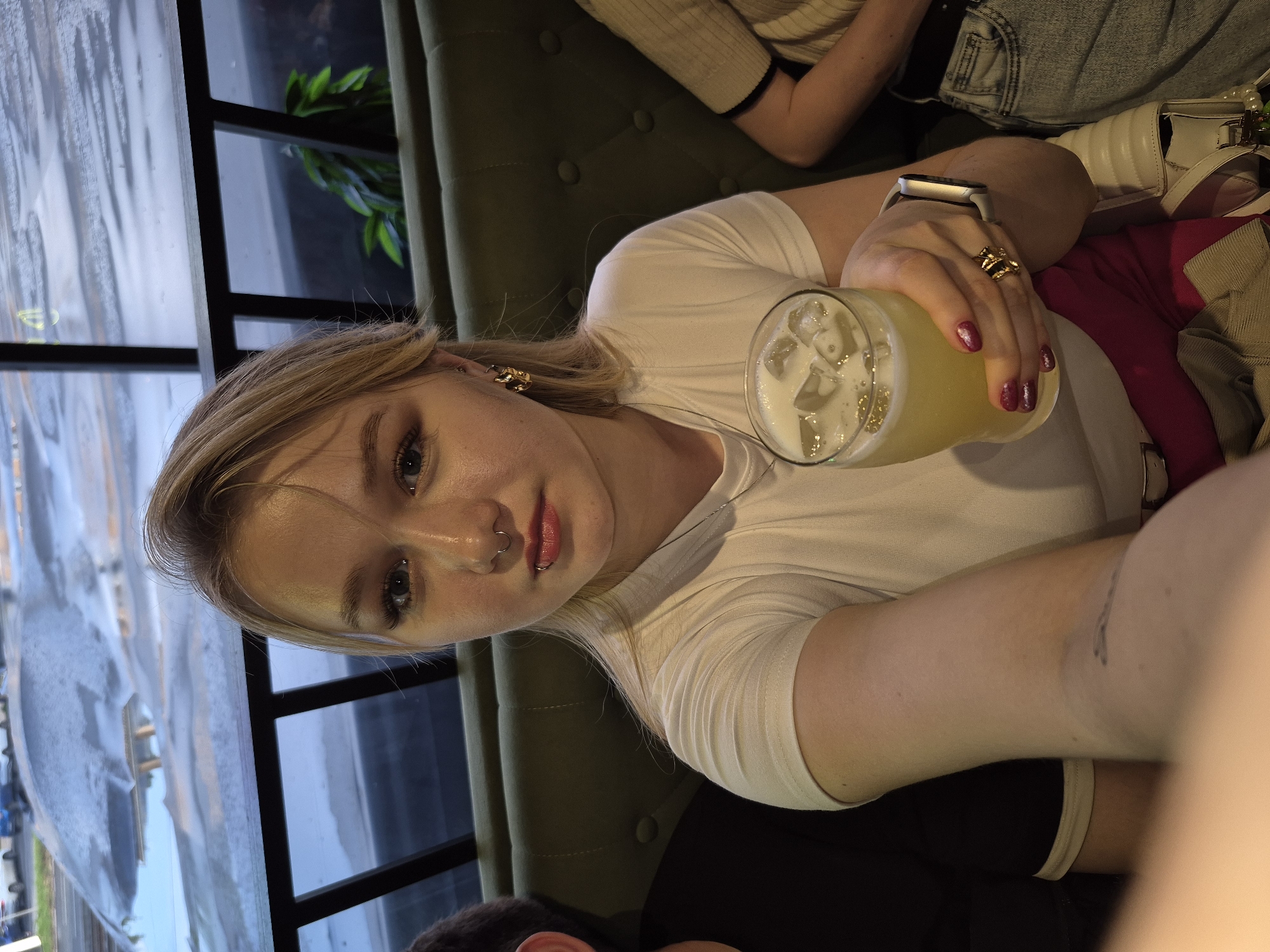 A young woman with blonde hair and a nose ring is holding a drink in a glass with ice. She is wearing a white t-shirt and has pink nail polish. To her left, a person with dark hair is visible, and to her right, another person wearing a beige top and jeans is seated. They appear to be in a dimly lit establishment with green upholstered seating. Through a window in the background, a street scene with cars and reflections on a wet surface can be seen, suggesting an urban environment, possibly in Izhevsk, Russia, given the context provided. The overall atmosphere suggests a casual outing at a restaurant or bar.
