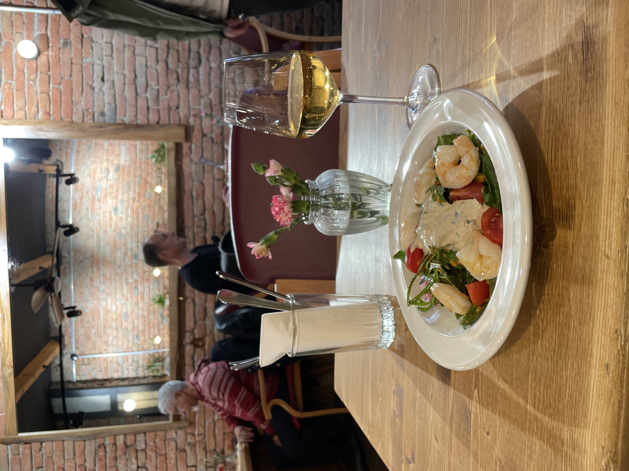 The image captures a dining scene in what appears to be a restaurant with a rustic interior. A wooden table dominates the foreground, displaying a plate of shrimp salad, a glass of white wine, and a small vase with pink flowers. Cutlery and napkins are visible in a glass holder. A reflection of a brick wall and people are visible in the background, suggesting the restaurant is located in a historical area. Based on the visual clues, the location could be Tbilisi, Georgia, given the prevalence of brick architecture and the common dining culture. People are present, but their actions are not clear from the cropped view.