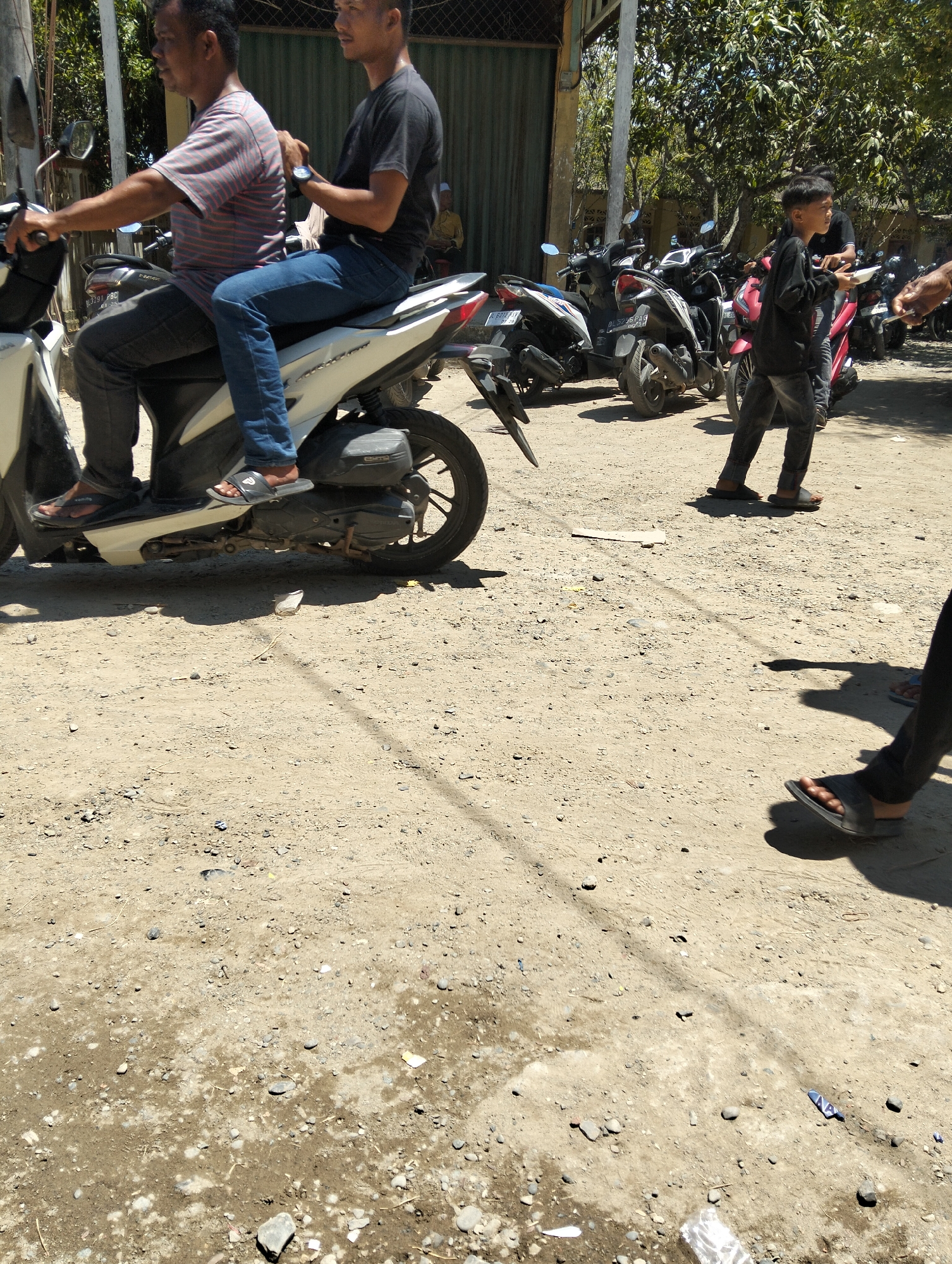 Two men are on a white scooter in a sunny, outdoor environment. The man in front is wearing a striped t-shirt and dark pants. The man behind him is wearing a dark t-shirt and blue jeans. The scooter is parked on a dirt and gravel surface. Several other scooters are parked behind the white scooter. In the background, there are trees and a building. A young boy in a black shirt and dark pants is standing to the right of the scooters, holding a white bowl. The overall scene appears to be in a village or a rural area.