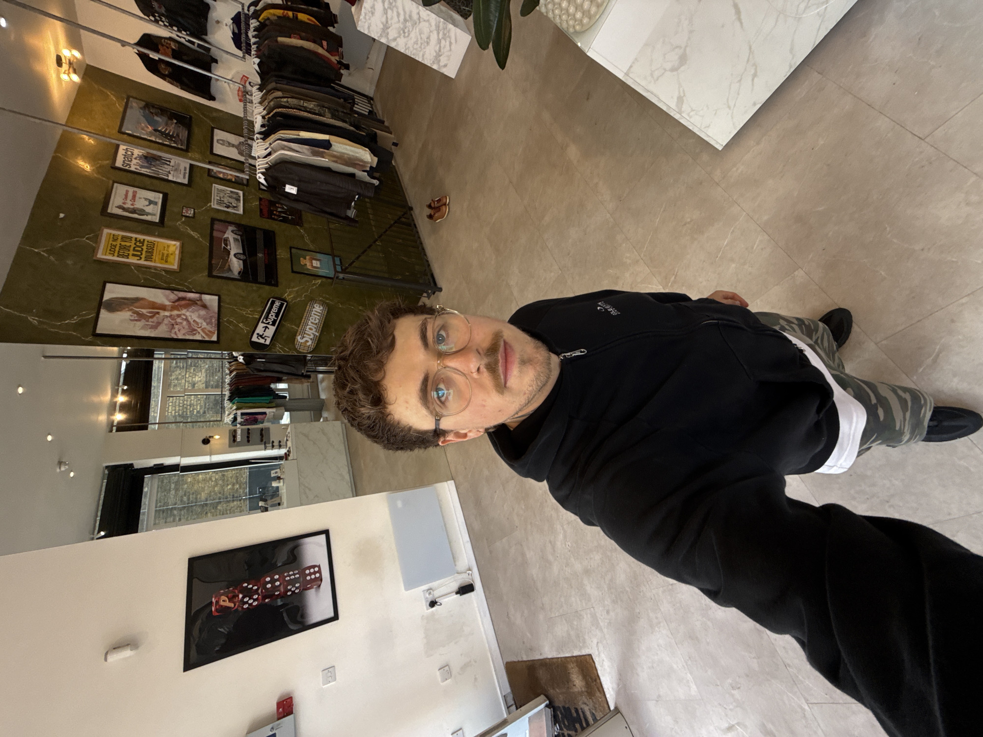A man wearing a black hoodie, camo pants and glasses is taking a selfie inside a clothing store. The store has a green wall with framed posters, clothing racks filled with various garments, and tiled flooring. A painting of dice hangs on a white wall. The store appears to be located in London, United Kingdom, based on the aesthetic and merchandise.