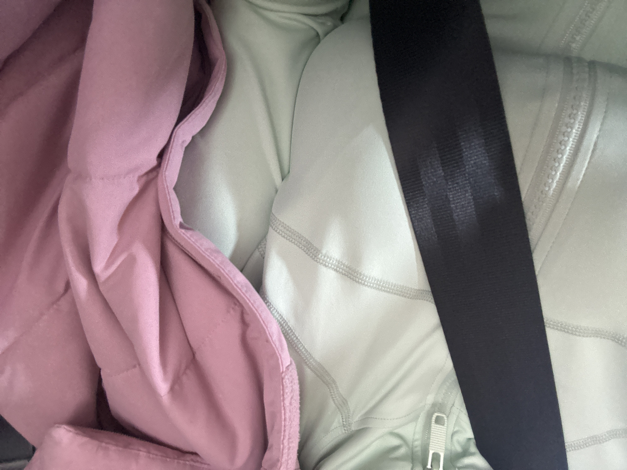 The image shows a close-up view of a black seatbelt restraining a pink puffer jacket and a pale green quilted jacket. The scene is confined within the vehicle, with no visible indication of the location being Moscow, Russia.