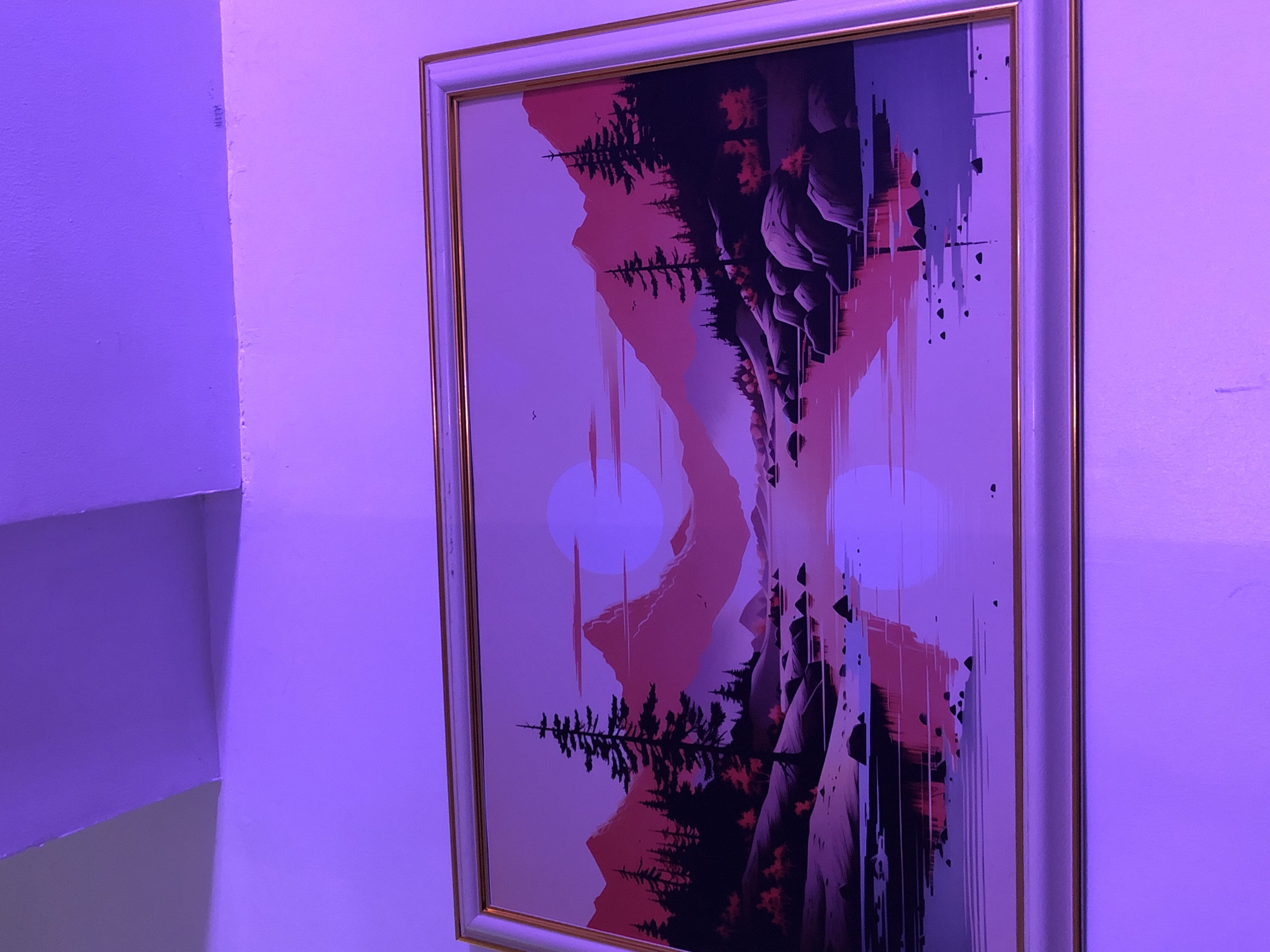 A framed painting hangs on a wall. The painting depicts a stylized landscape with mountains, trees, and two large circles resembling moons. The style of the painting incorporates a "glitch" effect with vertical lines and geometric shapes. The frame is white with a gold accent. The lighting in the room is predominantly purple, casting a violet hue on the wall and the painting. The wall is plain and slightly textured. Part of a wall corner is visible to the left.