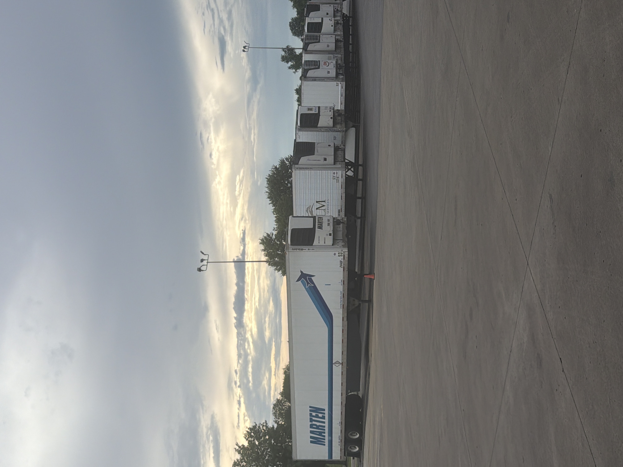 The image shows a line of refrigerated semi-trailer trucks parked in an asphalt lot. The trucks are white with varying logos and markings. One truck prominently displays "MARTEN" in large blue letters with a blue arrow graphic. Another truck has a logo with "LM SERVICES". The sky is overcast with some clouds, and there are trees visible in the background. A tall lamp post with multiple lights stands in the middle ground. The scene appears to be in a commercial or industrial area, possibly a truck depot or distribution center. The location is identified as Murfreesboro, United States of America.