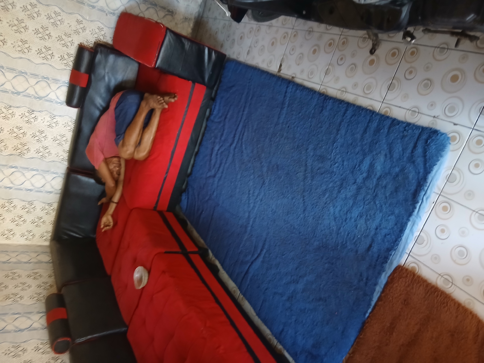 A person is sleeping on a red and black sectional sofa. The person is curled up on their side, wearing a pink shirt and blue shorts. A blue rug is placed on the floor in front of the sofa. A brown rug is also visible on the floor to the left of the blue rug. The room has tiled flooring and patterned wallpaper on the walls. A motorcycle is partially visible on the right side of the image. The scene appears to be indoors, possibly in a residential setting in Tenggarang, Indonesia.