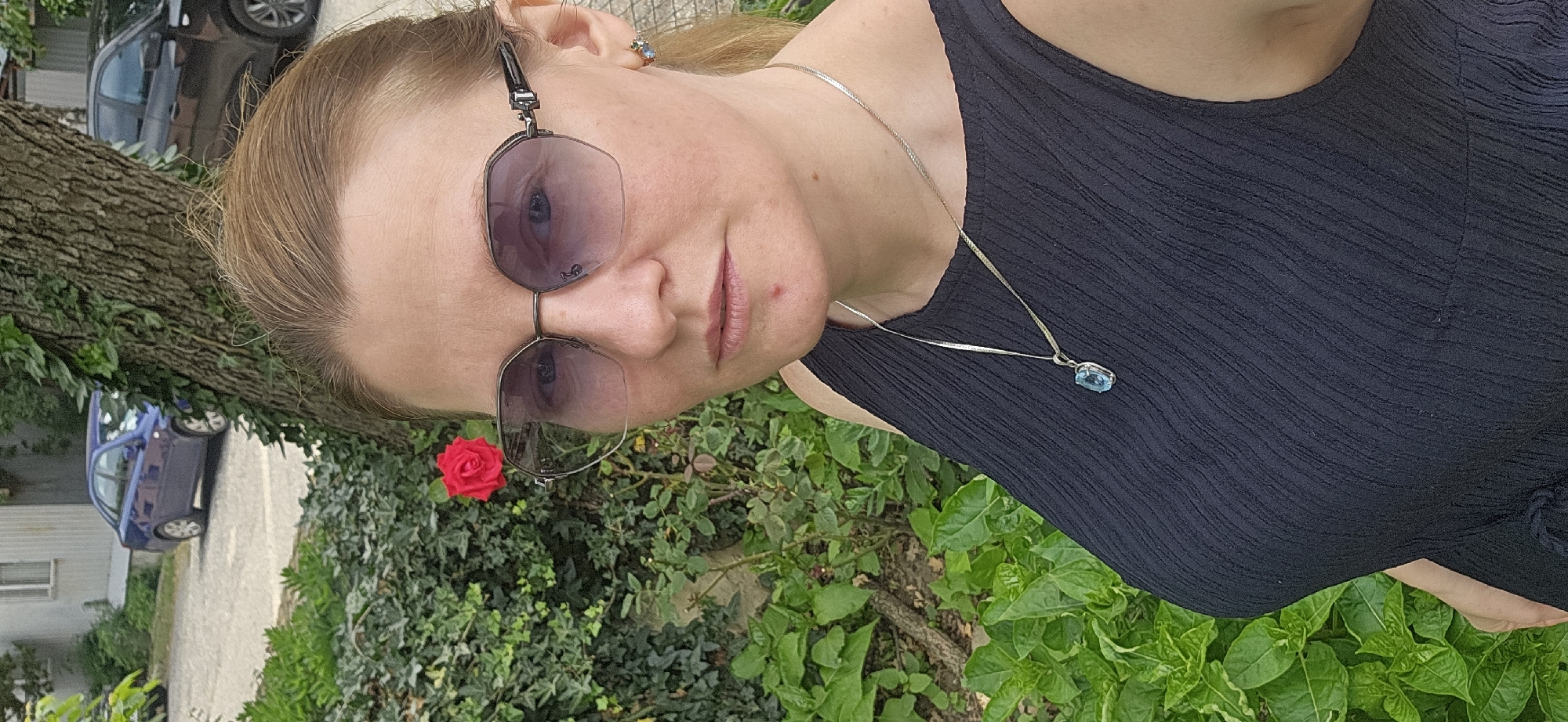 A middle-aged Caucasian woman is pictured in a close-up, outdoor shot. She is wearing sunglasses with a faceted frame, a black sleeveless top, and a silver necklace with a light blue pendant. Her hair is pulled back, and she has a small blemish on her chin. In the background, a red rose is visible on a bush. Further in the background, there are parked cars, including a blue sedan and a black SUV, on a paved area. Lush green foliage and a tree trunk are also present. The scene appears to be in a residential or park-like setting within the city of Dyurso, Russia.