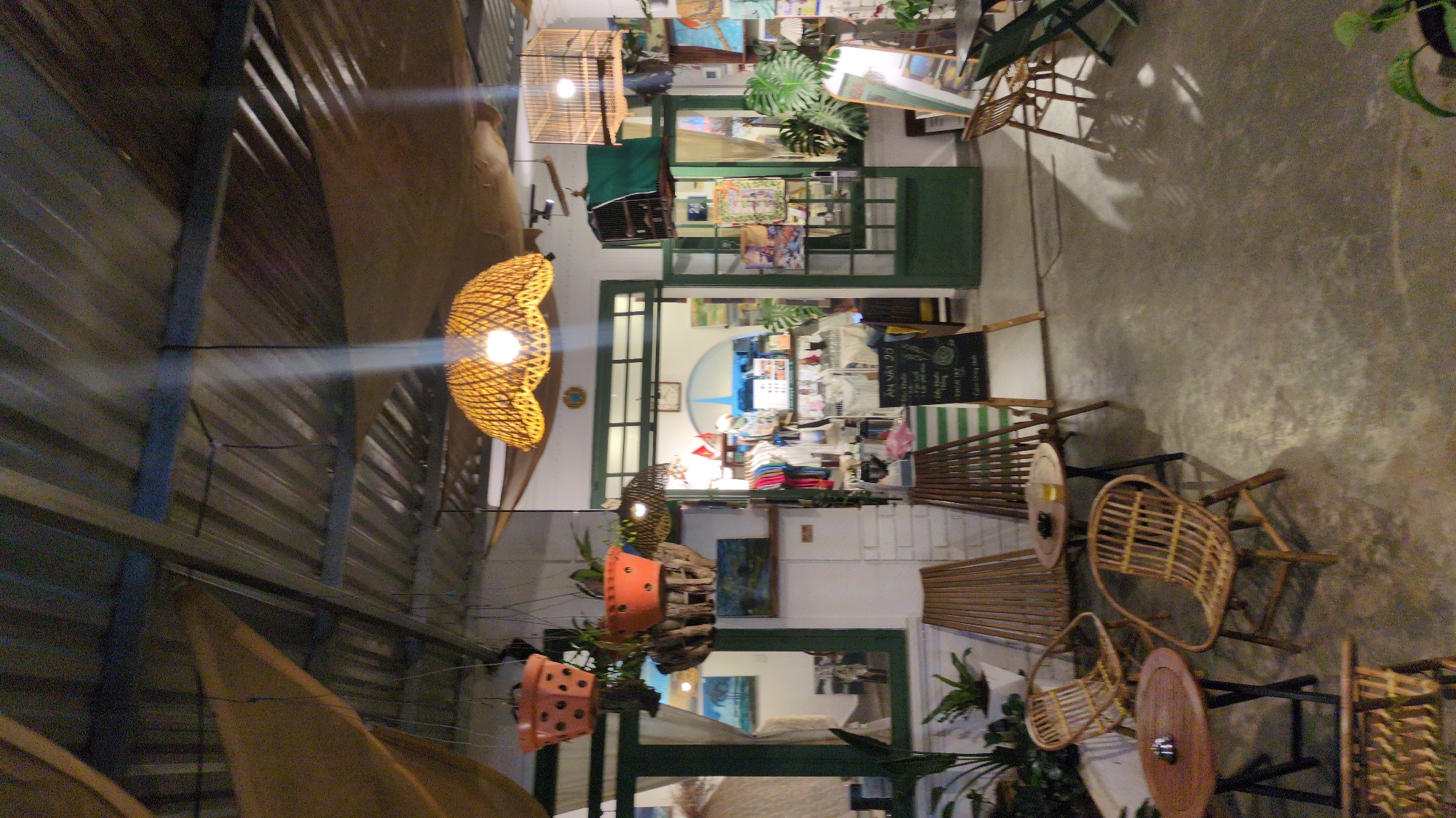 The image shows an indoor space in Cam Ly, Đà Lạt, Vietnam. The interior features a shop filled with various items, visible through a doorway with a green frame. The shop appears to sell clothing and other goods. Hanging plants in terracotta pots decorate the space, and multiple rattan chairs and tables are arranged in a seating area. Light fixtures with woven shades are suspended from the ceiling, casting a warm glow. The ceiling is corrugated metal.