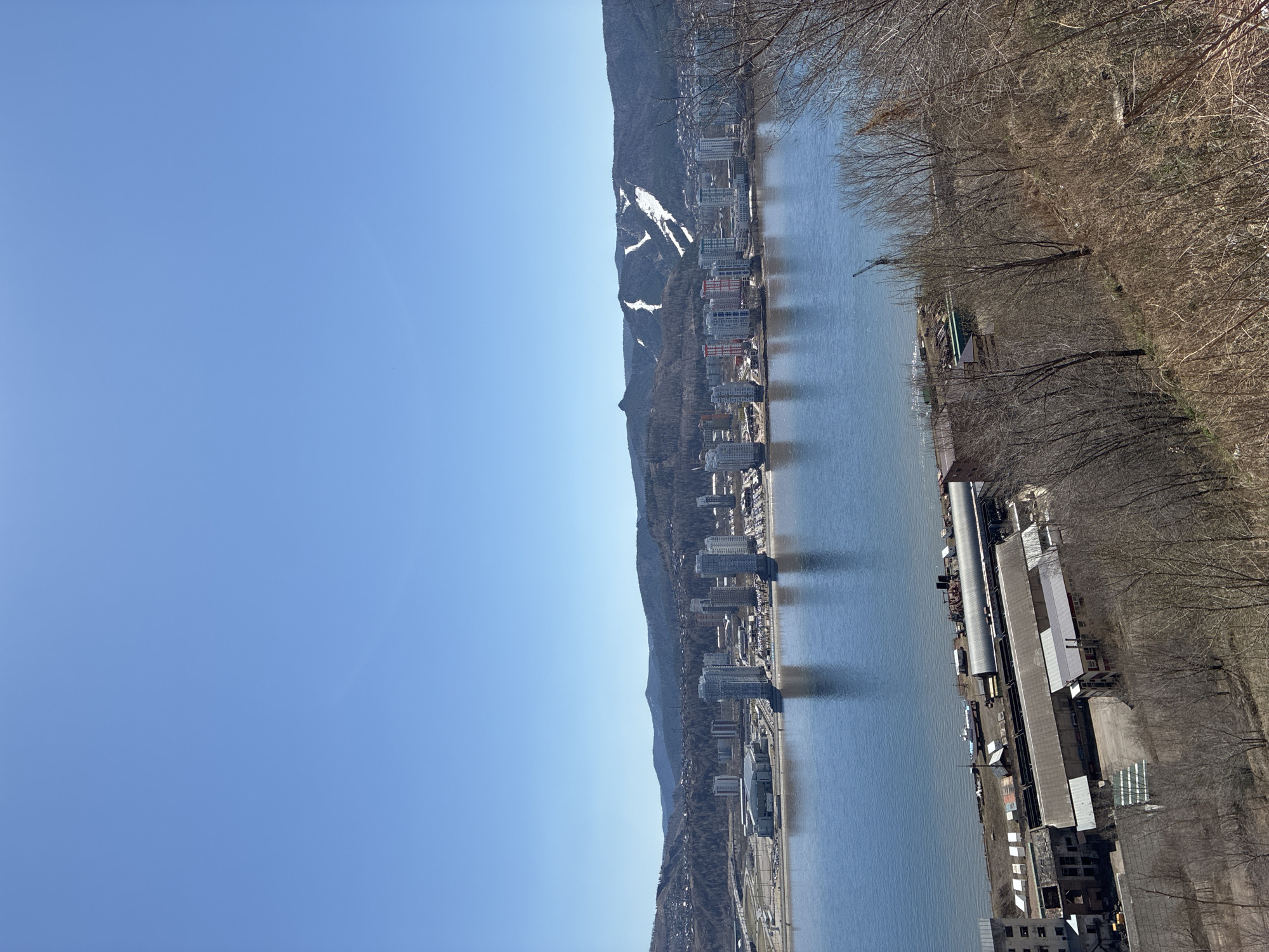 A vertical shot of a city situated along a river. The sky is a clear, pale blue, occupying a significant portion of the upper left quadrant of the image. The river bisects the image horizontally, reflecting the buildings on its banks. A mountain range with traces of snow stretches in the background. The riverbank on the right side of the frame is edged with bare trees and industrial buildings.