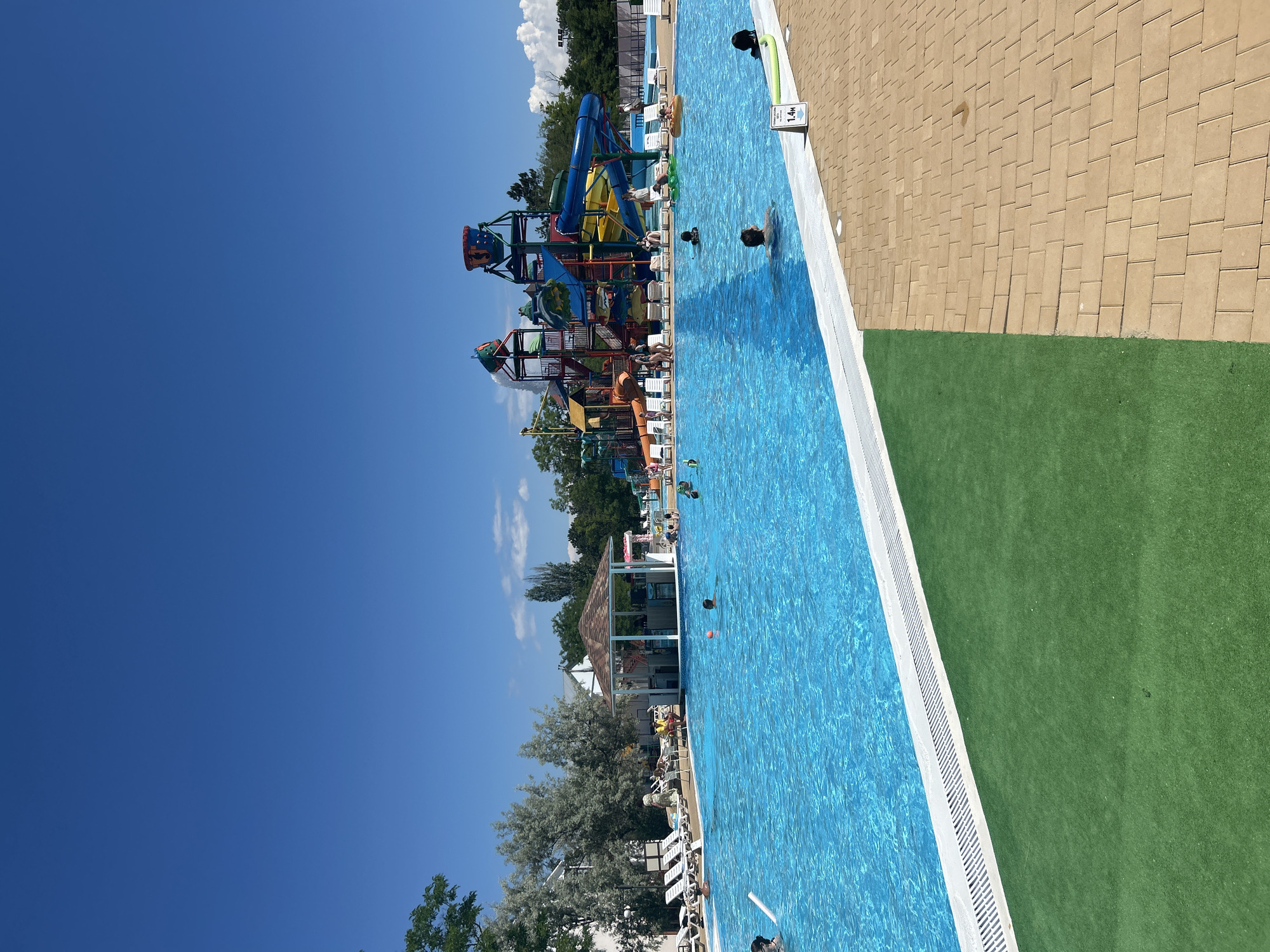 The image shows a bright, sunny day at a water park. In the foreground, a large, clear blue swimming pool takes up most of the frame. Several people are visible in the pool, some swimming and others floating on inflatable tubes. The pool area is lined with lounge chairs and umbrellas. In the background, a colorful water slide complex is the main attraction, featuring various slides and water play structures. A small, open-air bar or concession stand is situated near the pool. The environment is lush with green trees and vegetation surrounding the water park. The sky is a vivid blue with a few scattered white clouds. A sign indicating "1.4M" (likely depth) is visible near the edge of the pool. The scene appears to be a leisure activity area.