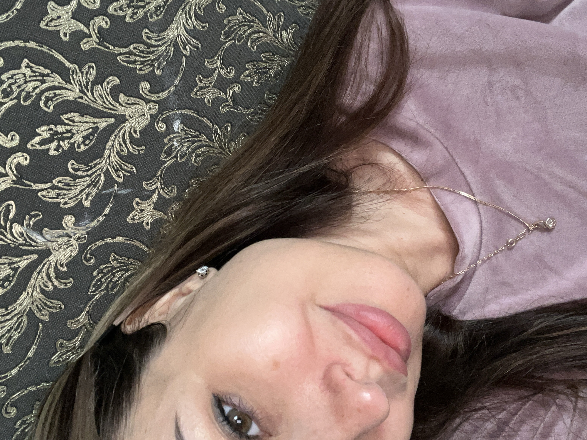 A close-up, waist-up, over-the-shoulder selfie taken by a woman with long, dark brown hair and brown eyes. She is wearing a light purple crew-neck shirt with a delicate gold chain and pendant necklace. Her face is turned towards the camera, with her left cheekbone and eye visible. She is wearing a small, diamond stud earring in her left ear. The background is a dark grey wall with a gold, damask-style pattern. The lighting is soft and diffused.