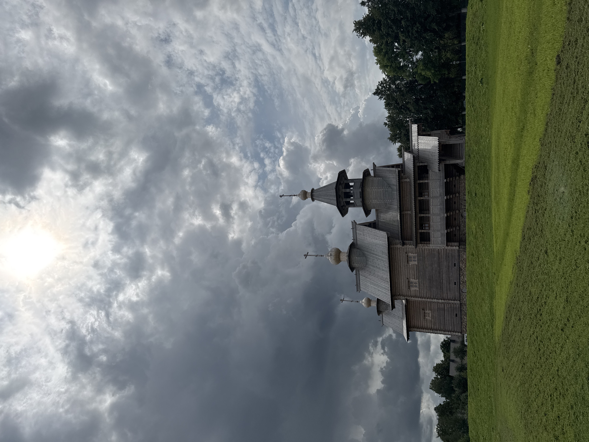 The image shows a wooden church with multiple domes and crosses, set against a dramatic sky with heavy clouds and bright sunlight filtering through. The church is constructed of logs and has a traditional architectural style, with a prominent bell tower. In the foreground, there is a large green grassy field, part of which appears to have been recently mown. Trees line the horizon behind the church. In the background, there are glimpses of other buildings and trees, suggesting a rural or semi-rural setting. The overall impression is one of a serene, yet imposing, religious structure in a natural landscape. This church is located in the village of Kamyanyuki in the Neman Rural Council, Belarus.