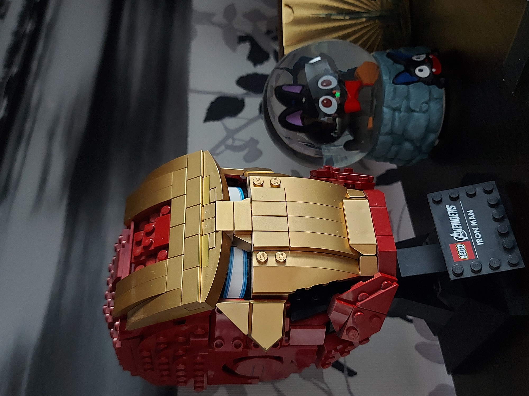The image shows a Lego Iron Man helmet positioned next to two small snow globes containing cartoon figures. The helmet is predominantly red and gold, with visible Lego construction. It rests on a black Lego stand labeled "LEGO Avengers IRON MAN." The snow globes are in the background, one containing a black cartoon cat with a red bow, and the other containing a cartoon figure with a blue cap. The items appear to be situated on a dark surface with a floral pattern. No location details are discernable.