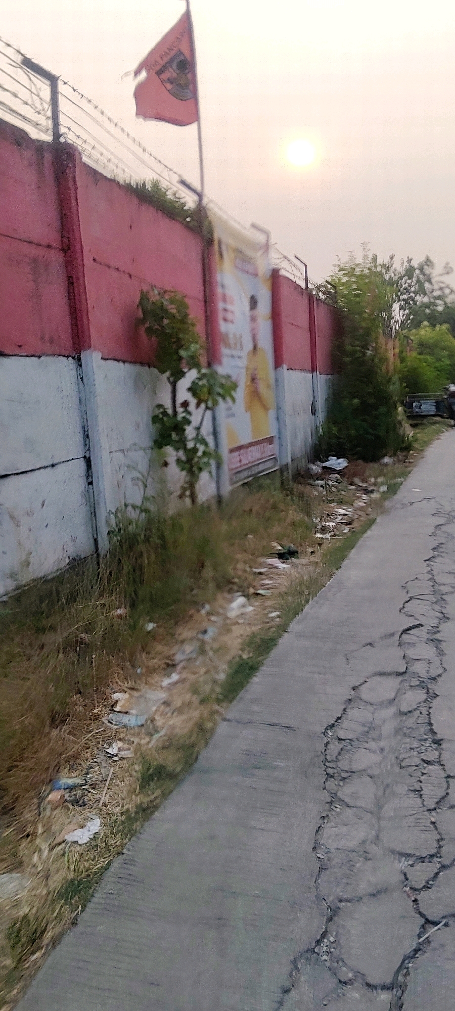 The image shows a red and white concrete wall with barbed wire on top. A red flag with a crest and text is flying on a flagpole. A large poster of a man in a yellow outfit is attached to the wall. There is a tree growing near the wall, and the ground is covered with dry grass and some litter. A paved road with cracks runs alongside the wall. The sky is hazy with a sun visible. This scene appears to be in the city of Mekarmukti, Indonesia.
