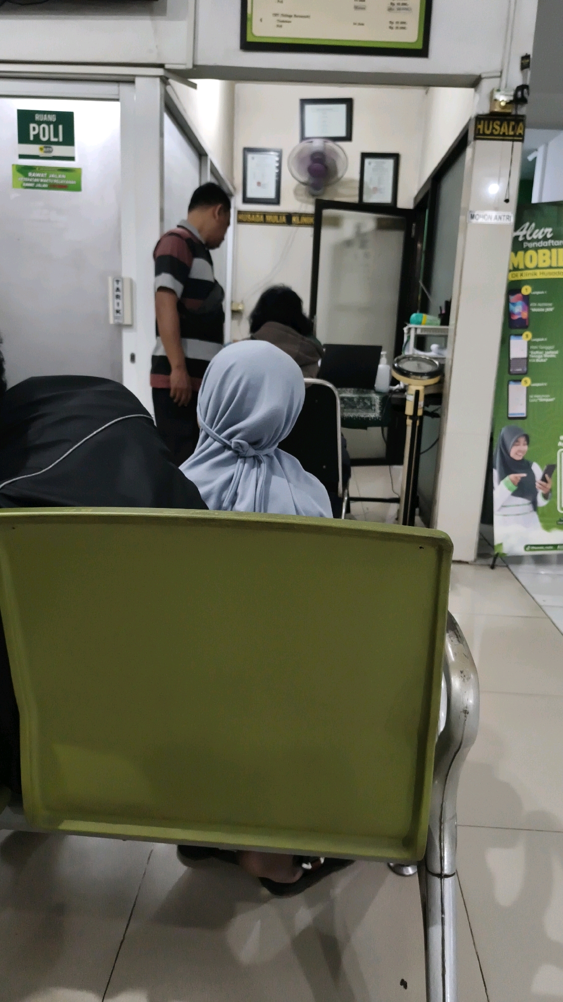 The image depicts the waiting area of a clinic, likely a polyclinic, in Rejoagung, Indonesia. The scene is dimly lit, with fluorescent lights overhead casting a sterile ambiance. In the foreground, a person wearing a light blue hijab is seated, their back to the camera. To their left, another person is partially visible, wearing dark clothing. In the mid-ground, a man in a striped shirt stands facing away from the camera, appearing to be in conversation with someone out of frame. A woman is seated at a desk in the background, possibly attending to administrative tasks. The clinic environment is suggested by a sign that reads "RUANG POLI" (Polyclinic Room) on a door to the left, and a sign that says "HUSADA" and "MOHON ANTRI" (Please Queue) to the right. Posters advertising healthcare services are also visible on the right wall. The overall impression is one of a busy, functional medical facility.