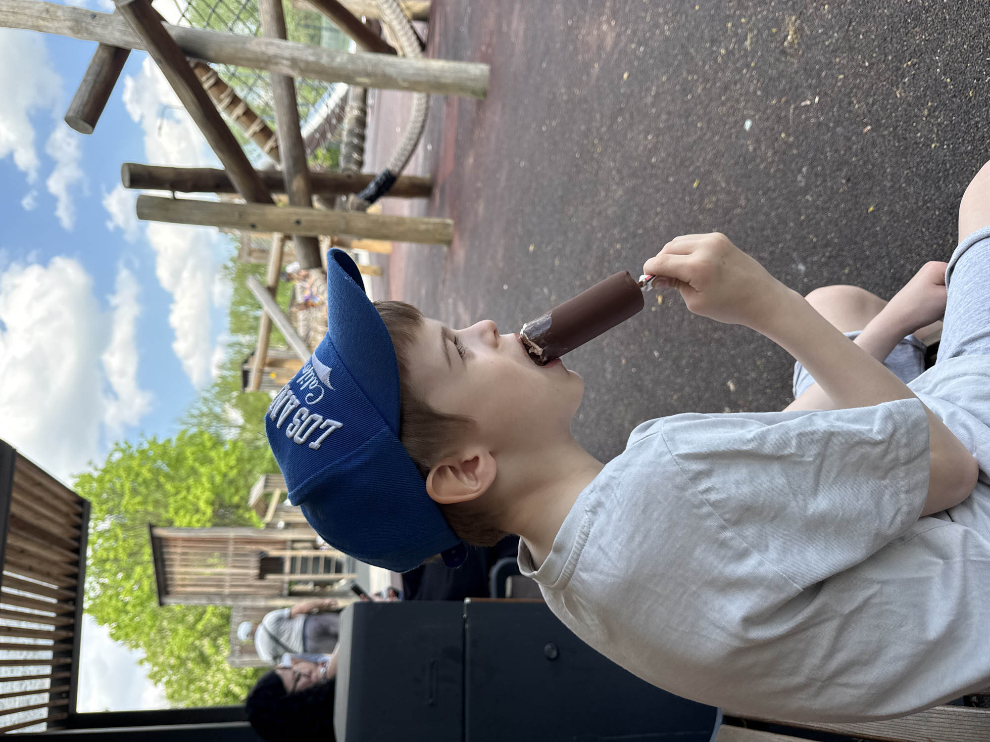 A boy wearing a blue baseball cap with the words "LOS ANGELES CALIF. 907" is eating a chocolate ice cream bar. He is wearing a gray t-shirt and gray shorts. The setting appears to be a playground with wooden structures. The location is not Moscow, Russia.