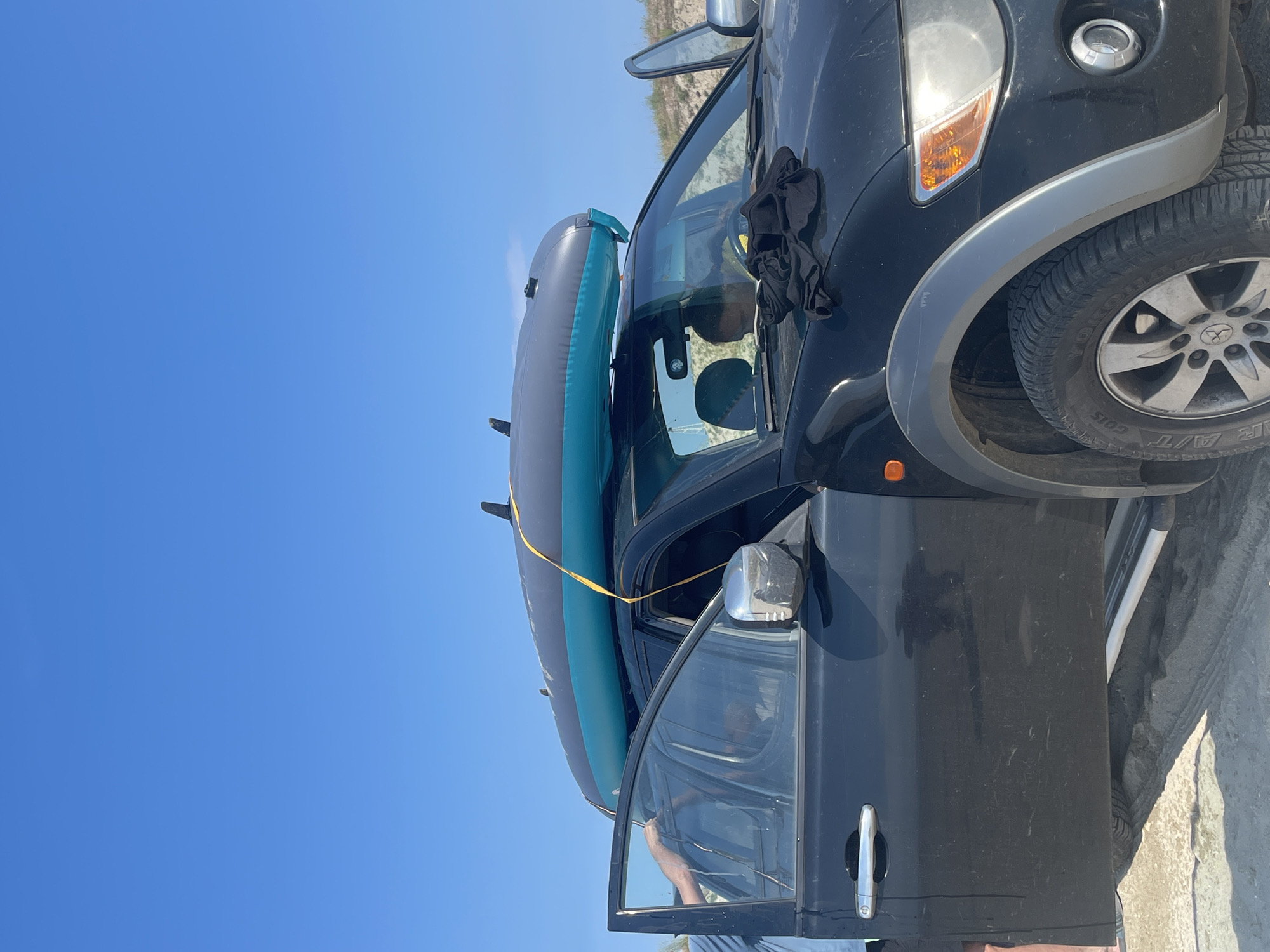 An eye-level, vertical, outdoor shot shows a black, four-door pickup truck with its front passenger door open, parked on a sandy surface under a clear blue sky. The truck's roof rack is equipped with a deflated, grey and teal inflatable kayak, secured with a yellow strap. A person's arm and shoulder are visible through the open driver's side door. A black t-shirt is draped over the front of the truck's hood. The location is Agios Georgios, Greece.