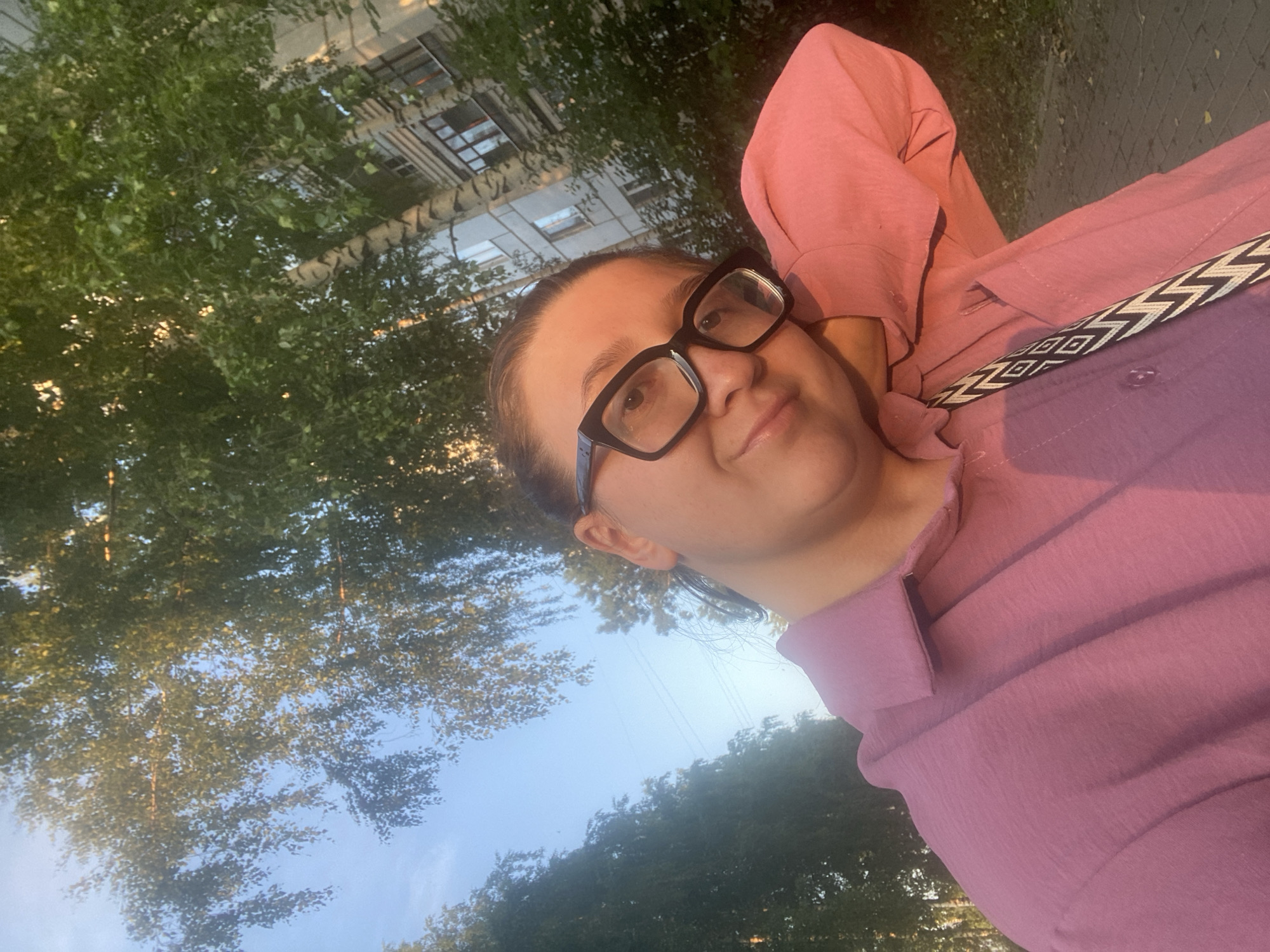 A woman with dark hair pulled back and wearing large black-framed glasses poses for a selfie outdoors in Chaikovsky, Russia. She is dressed in a pink collared shirt and a patterned strap crosses her chest. The background features trees with lush green leaves and a building with multiple windows, characteristic of urban residential architecture. The lighting suggests it is either early morning or late afternoon, with the sun casting a warm glow on the scene. The pavement in the foreground indicates a sidewalk or path.