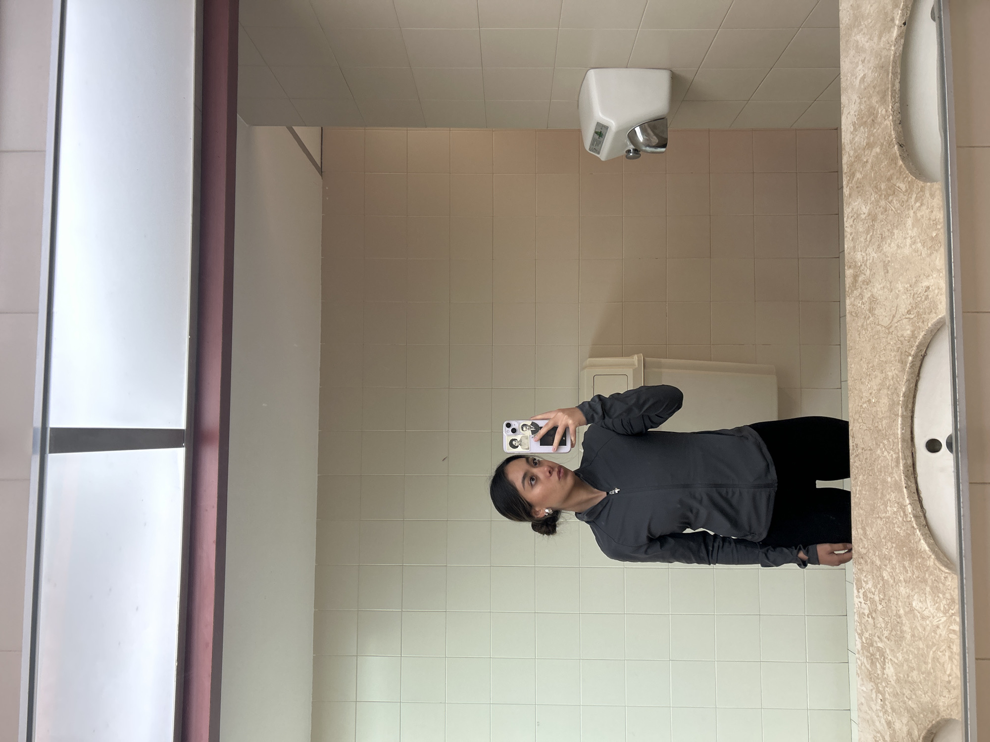 A woman is taking a mirror selfie in what appears to be a public restroom or bathroom. She is positioned in the lower-middle portion of the mirror's reflection, holding a smartphone with a light purple case and stylized portraits on the back. Her gaze is directed towards the phone screen. She has dark hair pulled back into a bun and is wearing a dark grey, long-sleeved, zip-up athletic jacket and black pants. Silver earrings are visible on her ears.
The room reflected in the mirror features light beige or off-white square tiles on the walls and a lighter, grid-patterned ceiling. A white hand dryer is mounted on the wall in the upper right quadrant of the reflection. Below the hand dryer, a white rectangular object, possibly a paper towel dispenser or part of a toilet, is partially visible. To the right of the woman, a textured, light brown and white speckled countertop or sink area is visible, with a white basin partially shown on the far right edge of the mirror. A narrow, dark pink or maroon frame runs along the left edge of the mirror, adjacent to a white panel which reflects a window frame with horizontal dividers. The overall lighting suggests an indoor setting with artificial light. No legible text is clearly visible.