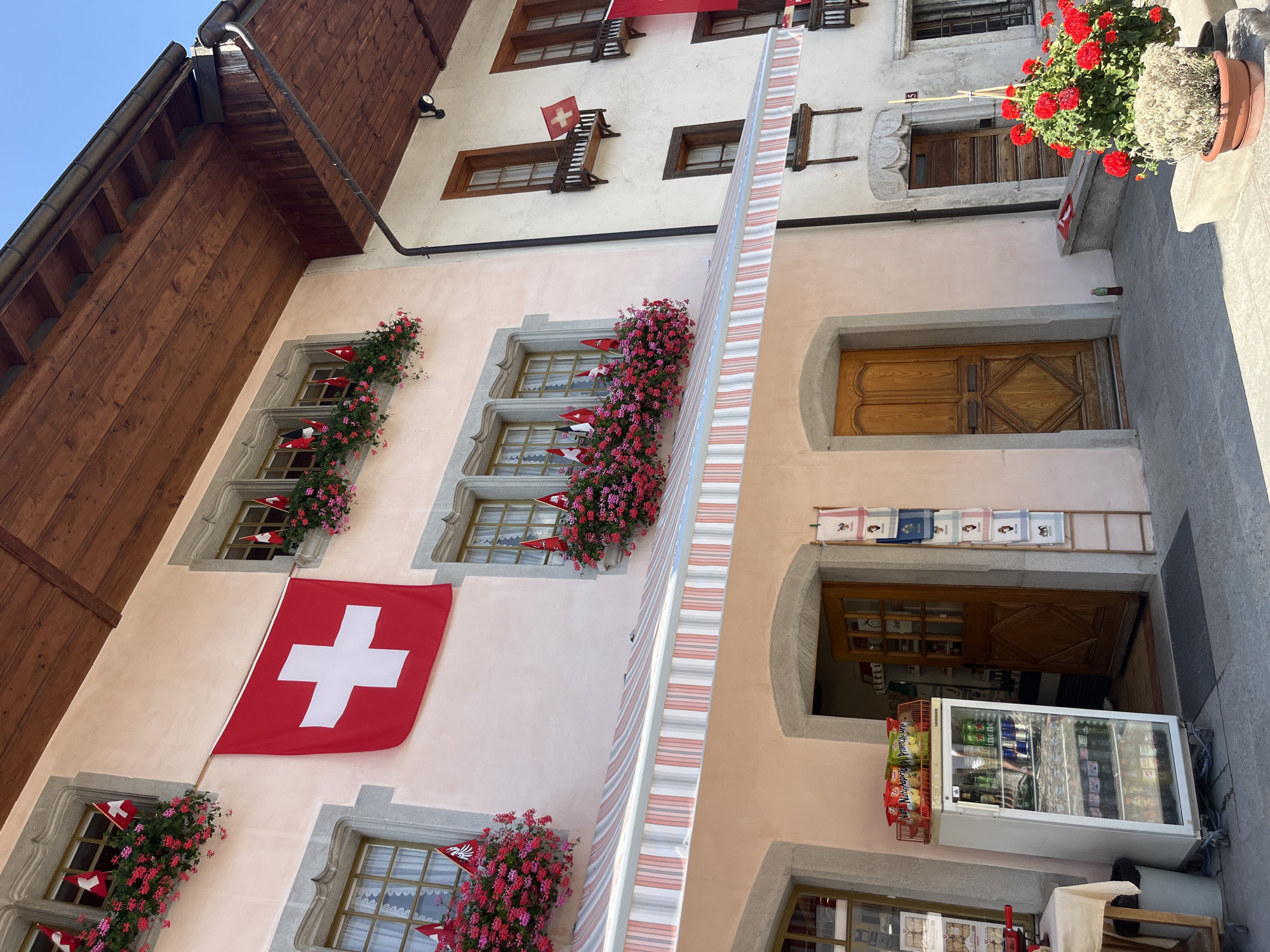 This is a ground-level, eye-level, medium shot of a street scene in Epagny, Switzerland. The building in the foreground is painted light pink and features several windows adorned with floral arrangements and Swiss flags. A large Swiss flag is prominently displayed on the facade. An awning with pink and white stripes extends over the entrance to a shop. Inside the shop, a refrigerator is stocked with beverages. Next to the shop, a wooden door is visible. To the right of the pink building, a white building features windows with small balconies displaying Swiss flags and more floral arrangements. A red Swiss flag is also visible on this building. Red flowers in a pot are situated in front of the white building. The scene is bathed in sunlight, and the sky is clear.