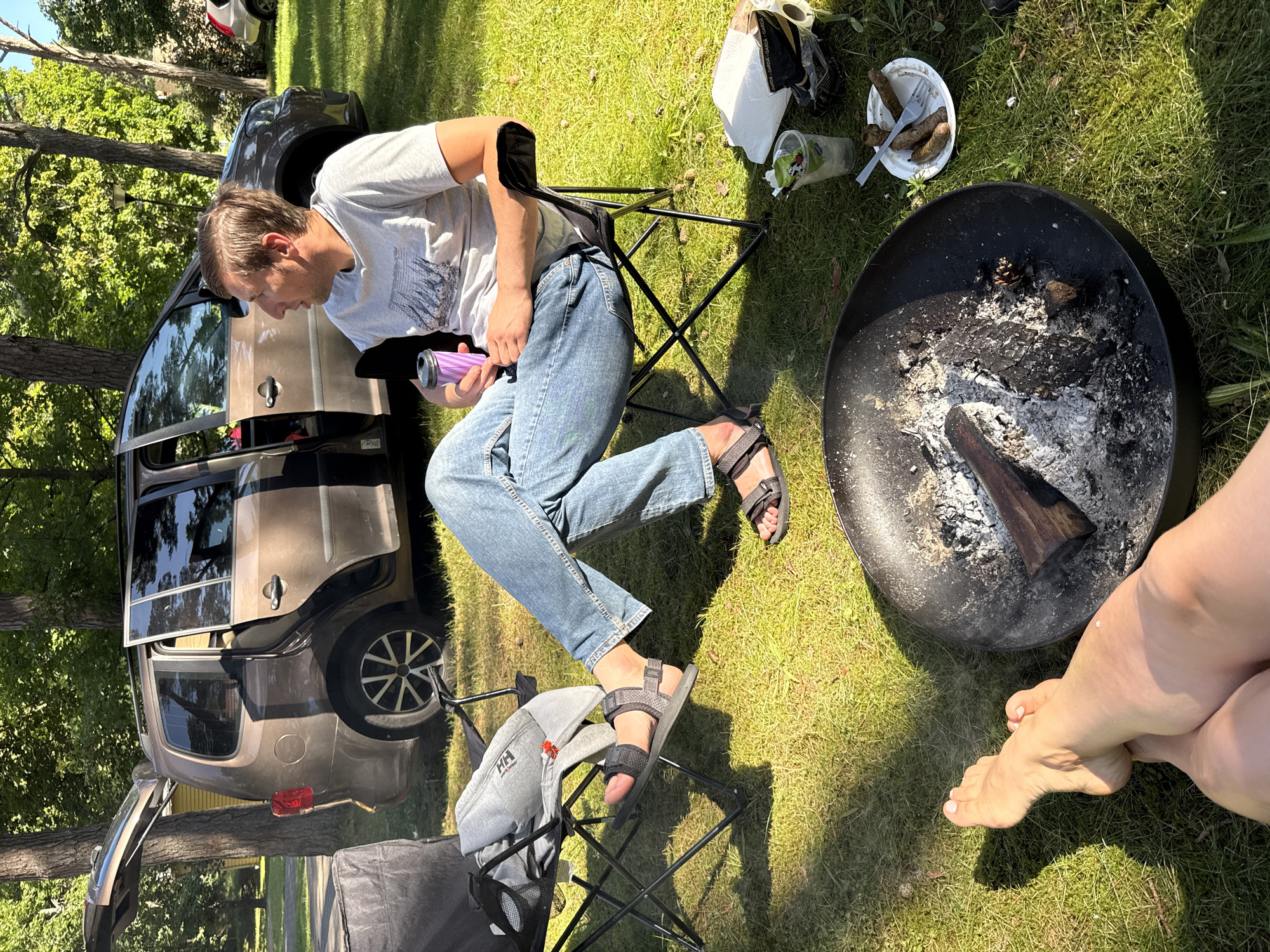 The image depicts an outdoor scene likely at a campsite or park. In the background, a tan minivan with its rear hatch and driver's side door open is parked on a grassy area with trees. In the foreground, a man is seated in a black folding chair, holding a purple tumbler. He is wearing a grey t-shirt and blue jeans. In front of him, another black folding chair has a grey hooded sweatshirt with "HH" logo draped over it. To the right of the man, a fire pit with ashes and a piece of wood is visible. A white paper plate with three sausages and a fork, a plastic cup with a beverage and a straw, and a roll of toilet paper are scattered on the grass nearby. The lower part of the image shows a person's bare legs and feet, suggesting another individual is present and relaxing. The overall environment is a sun-drenched grassy area with scattered pinecones, characteristic of an outdoor recreational space. There is no specific indicator within the image to suggest a location in Ventspils, Latvia.