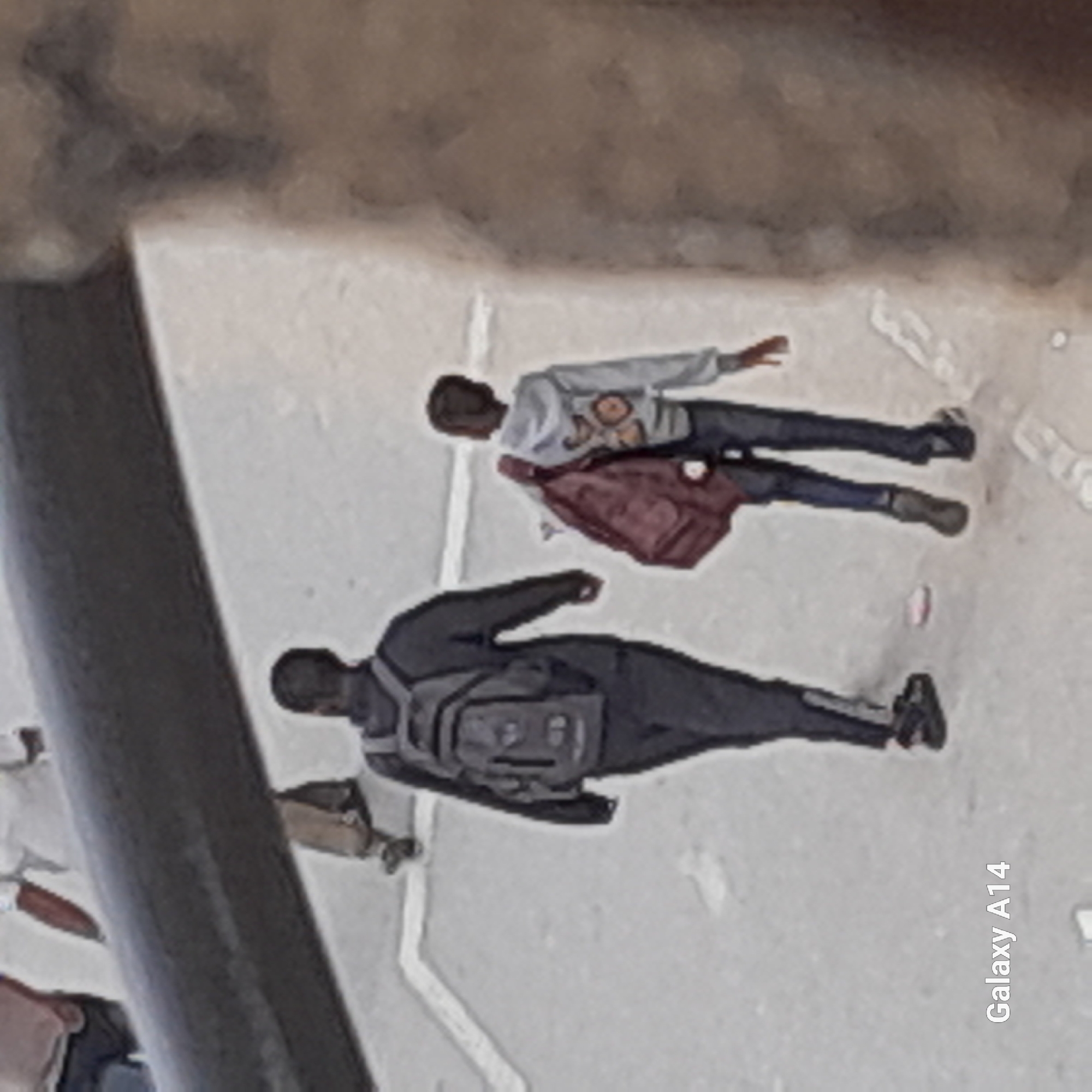 Two individuals are walking on a street in an urban setting. The first individual is wearing dark clothing and a backpack. The second individual is wearing a lighter-colored top, dark pants, and carrying a red bag. The scene appears to be taken from an elevated perspective. The location is identified as "Unknown, Unknown" in the image.