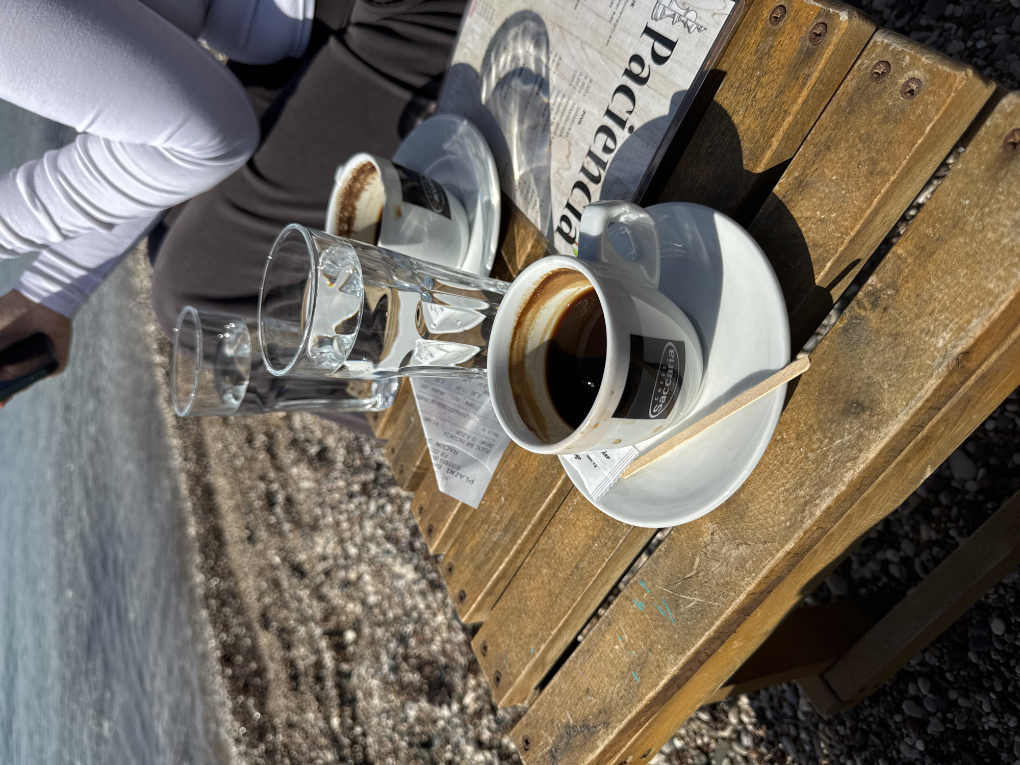 The image depicts a wooden table at a cafe near a beach. The table is set with two cups of coffee, two glasses of water, and a menu. The coffee cups have the "Cafe Saccaria" logo. The menu is for "Paciencia Montenegro". A receipt is visible on the table. A person is partially visible in the background, wearing white pants. The beach is composed of small pebbles and the sea is visible in the background. Given the "Paciencia Montenegro" and "Racun" in the OCR, this scene is in Montenegro. Given "Paciencia BAR", it is highly likely that the image depicts a scene at the Paciencia bar in Budva, Montenegro.