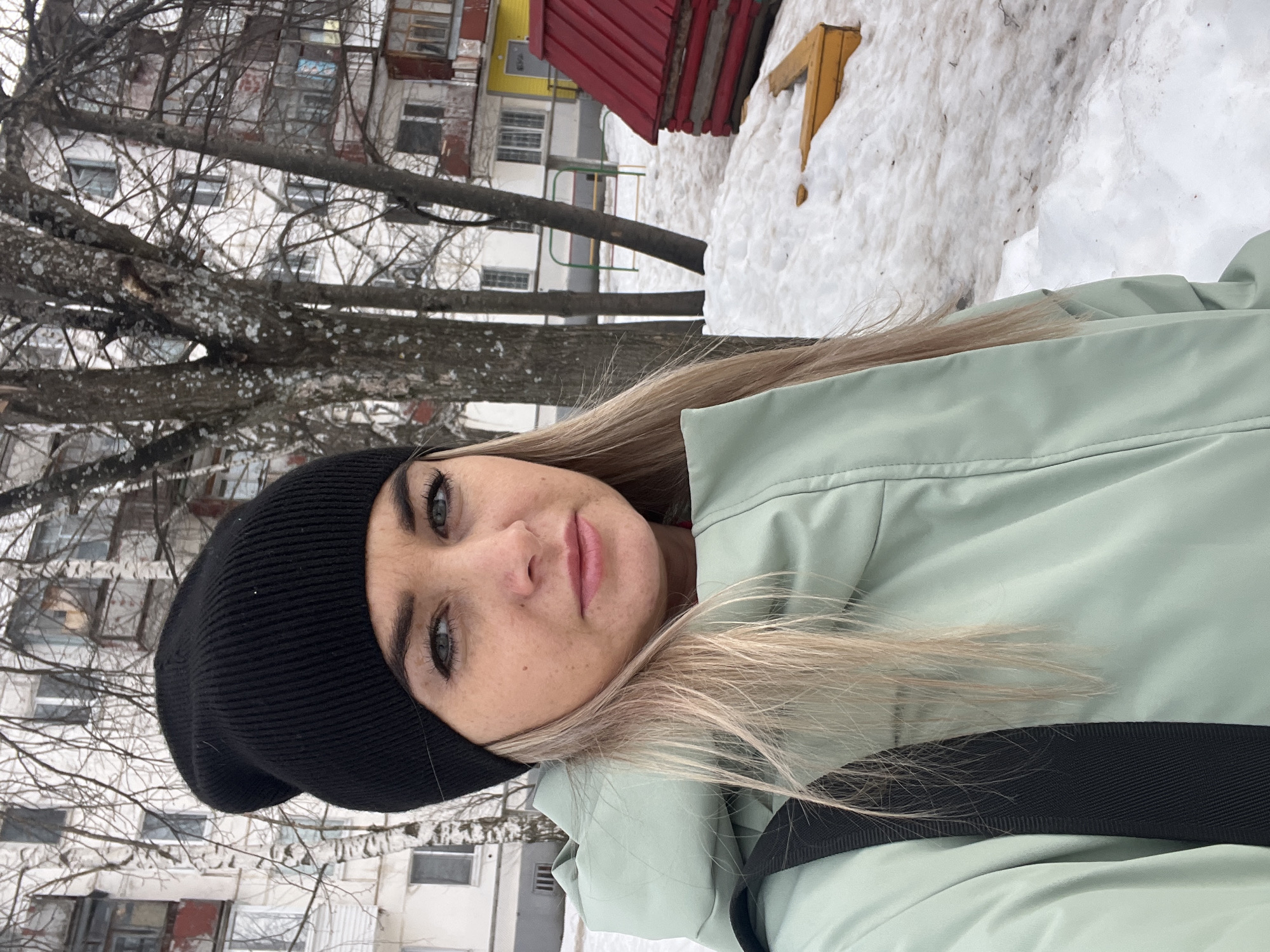 A blonde-haired woman with light eyes, wearing a black knit beanie and a mint green puffer jacket with a visible black strap, is captured in a waist-up, direct-gaze selfie outdoors in Chaikovsky, Russia. The ground is partially covered with melting snow. Bare deciduous trees and multi-story residential buildings with various windows and balconies are present in the background. Playground equipment, including a red-roofed wooden play structure, yellow wooden sandbox edging, and a green and yellow metal climbing frame, is visible on the right, partially obscured by trees and located near a building with a yellow-painted lower section. The scene indicates a late winter or early spring season.