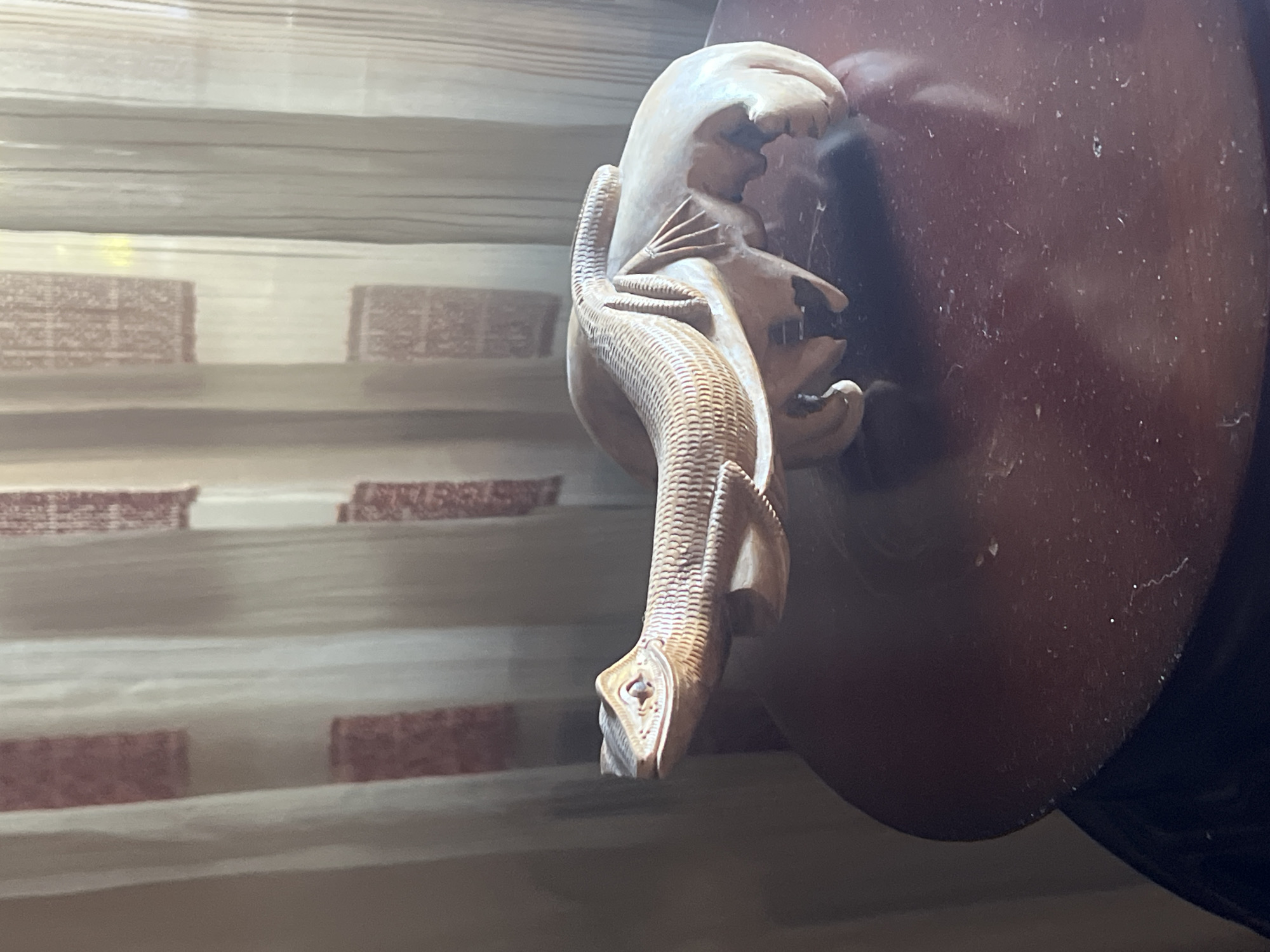 A wooden sculpture of a lizard is displayed on a dark, polished wooden table. The lizard, carved with intricate scales, rests on a larger, unfinished wooden form that resembles a rock or a natural element. The sculpture is positioned in front of a sheer, cream-colored curtain with a repeating pattern of reddish-brown squares. The lighting suggests it is indoors, possibly in a home or a shop. The overall scene is still and quiet.
