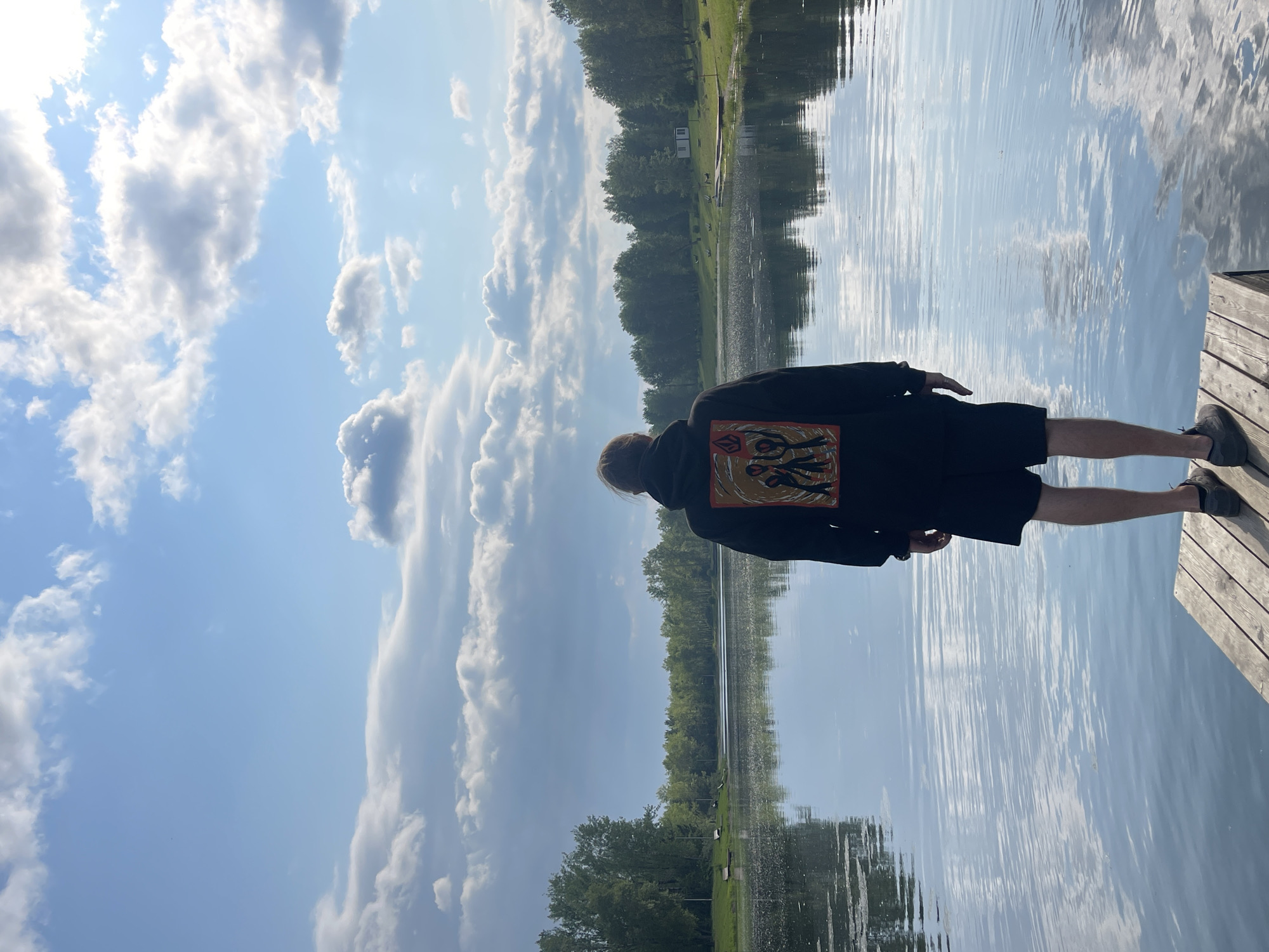 A person stands on a wooden dock facing a calm lake. The person is wearing a black hoodie with a graphic design on the back and black shorts. The lake is surrounded by trees and a green lawn. The sky is blue with scattered white clouds. The scene appears to be a park or a recreational area. The location is the city of Krasnoyarsk, Russia.