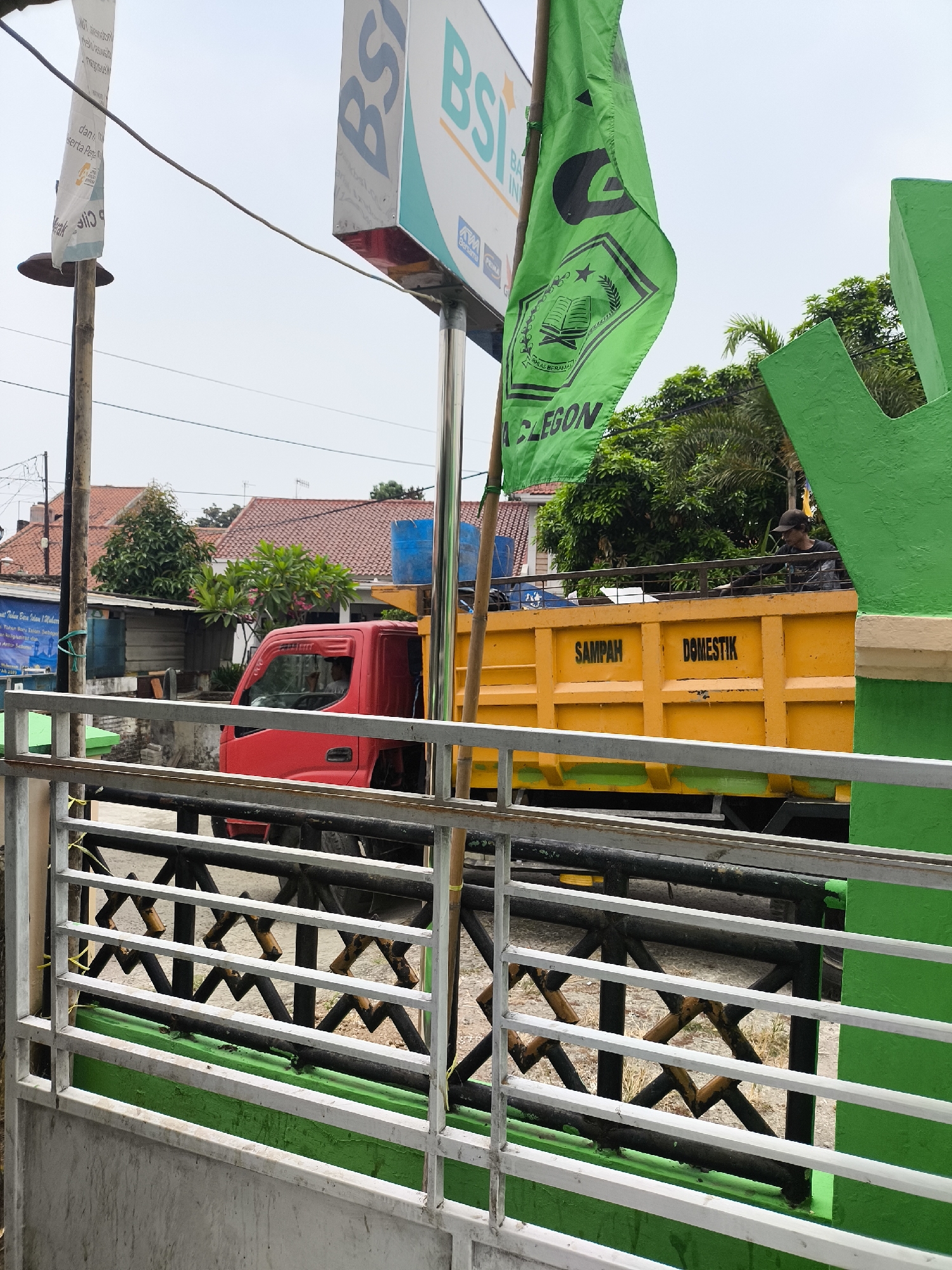 The image depicts a street scene in Cilegon, Indonesia, characterized by a bright red truck with a yellow dump bed positioned behind a metal fence. The dump bed of the truck has the words "SAMPAH DOMESTIK" (Domestic Waste) printed on it. A person wearing a dark shirt and a cap is visible in the truck's cab. In the background, residential buildings with red tile roofs are visible, along with trees and a blue water tank. A green flag with black lettering and a circular emblem is prominently displayed, waving from a pole. To the left of the flag is a sign that reads "BSI" (Bank Syariah Indonesia), with smaller text below it. The overall environment suggests a typical urban or semi-urban setting in Indonesia.
