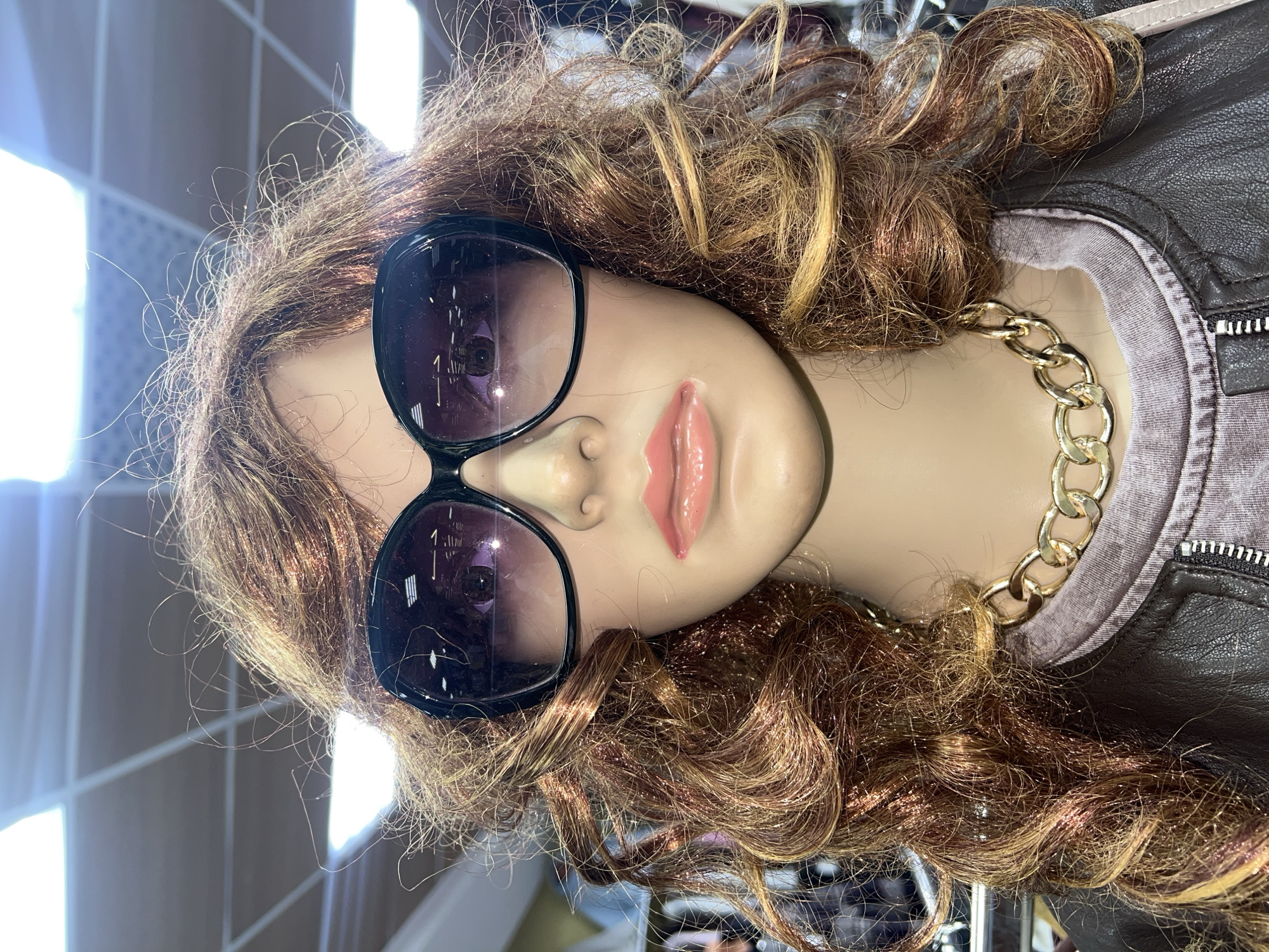 This image displays a mannequin's head and upper torso, presented in an indoor retail setting in Perm, Russia. The mannequin is positioned as if lying on its left side, with its head slightly tilted.
The primary subject is the mannequin, which features long, voluminous, curly hair styled in a mix of brown and blonde shades, resembling a wig. The hair appears somewhat tousled with individual strands visible, particularly at the top and around the face. The mannequin is wearing large, black, oversized sunglasses that obscure its eyes. Its lips are painted with a glossy pink lipstick and are slightly parted.
Around the mannequin's neck is a prominent, thick gold-colored chain necklace. Beneath the necklace, a plain light grey or light purple crew-neck top is visible, layered under a dark brown jacket. The jacket appears to be made of leather or a similar textured material, with visible stitching and a silver-colored zipper detail on the right side of the frame.
Reflections in the sunglasses reveal elements of the indoor environment, including bright light sources and some architectural lines, suggesting overhead lighting or windows within the commercial space. The background is softly blurred, reinforcing the impression of an indoor display area. There is no activity taking place, as it is a static retail display. No visible text is present in the image.