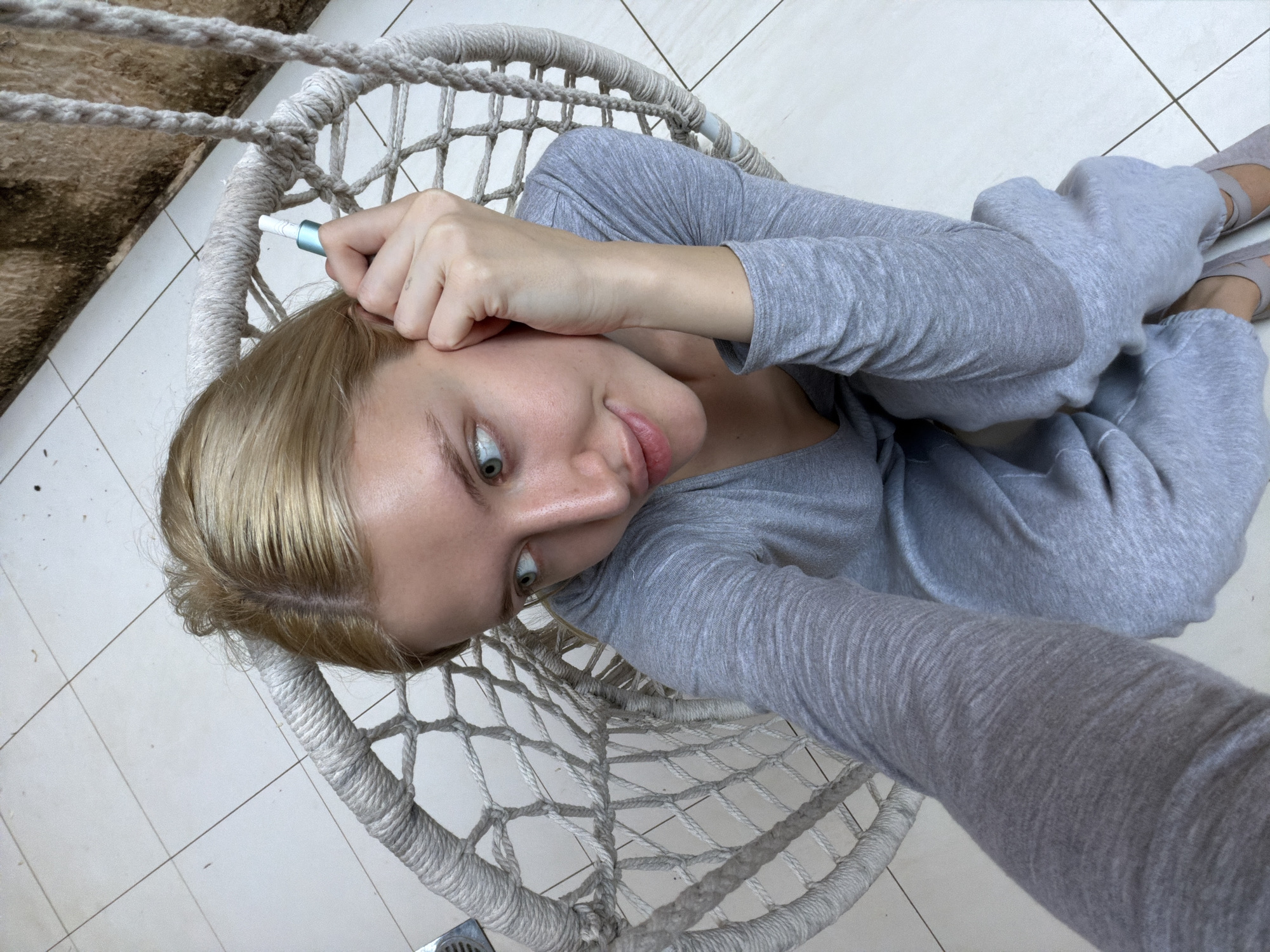 A woman with blonde hair and light gray clothing is sitting in a white rope chair. She is holding a light blue object near her head. The background appears to be a tiled floor. The location is Nerin, Montenegro.