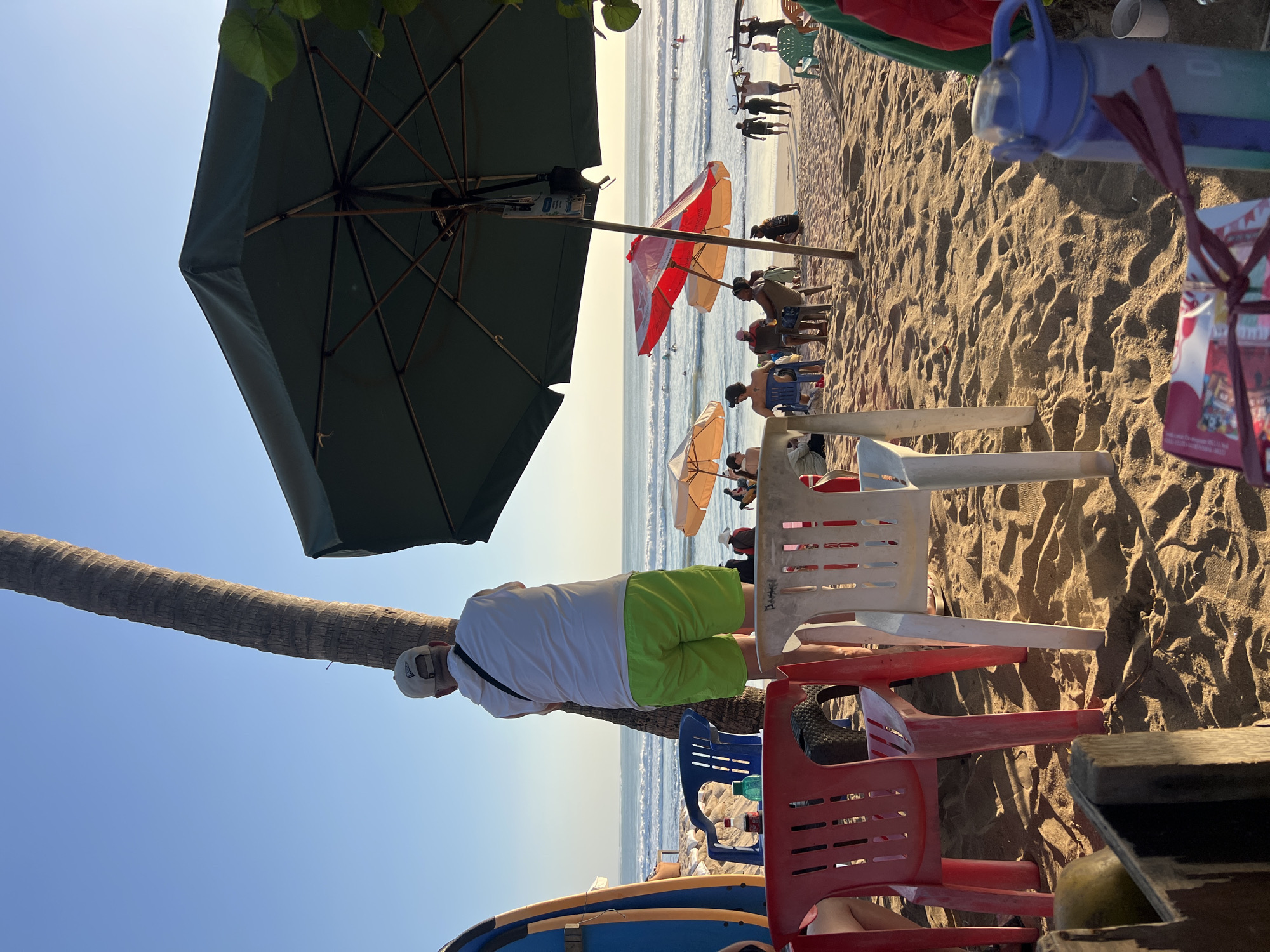 A man wearing a white t-shirt and bright green shorts stands with his back to the camera, leaning against a palm tree on the sandy beach of Kuta, Indonesia. The ocean with gentle waves is visible in the background, with surfers in the water. Several beach umbrellas, one large dark green and another red and white, are set up on the sand, providing shade for groups of people seated on chairs. Colorful plastic chairs, a blue surfboard, and a water bottle are scattered on the sand in the foreground. The sun is setting, casting a warm golden light across the scene.