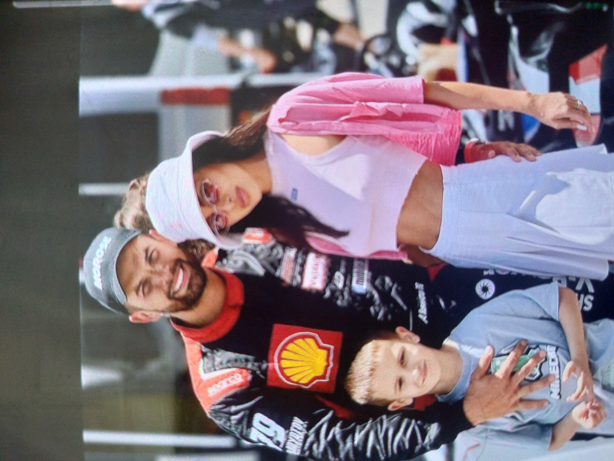 The image shows a smiling man in a black and red racing suit with a Shell logo, wearing a grey cap. He has his arm around a young boy in a grey t-shirt. Beside the man is a woman wearing a white crop top, a pink jacket, and a white cap. The background is blurred, suggesting an outdoor event, but the specific location in Tsarevo, Bulgaria cannot be determined from the image.