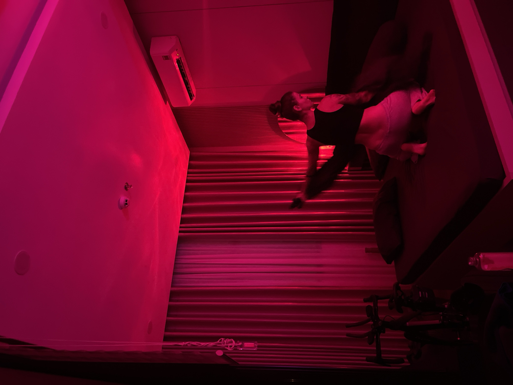 A dimly lit room, bathed in red light, features a person kneeling on a bed. The person is wearing a black top and light-colored bottoms, with their hair in a bun. They appear to be in the process of moving, with one leg extended. To the left of the bed is a stationary exercise bike. In the background, red curtains cover a window, and an air conditioning unit is mounted on the wall. A small mirror is visible on the wall behind the bed. The overall scene suggests a private space, possibly a bedroom or a personal gym area. The mention of Đà Nẵng, Vietnam, provides a geographical context for the location.