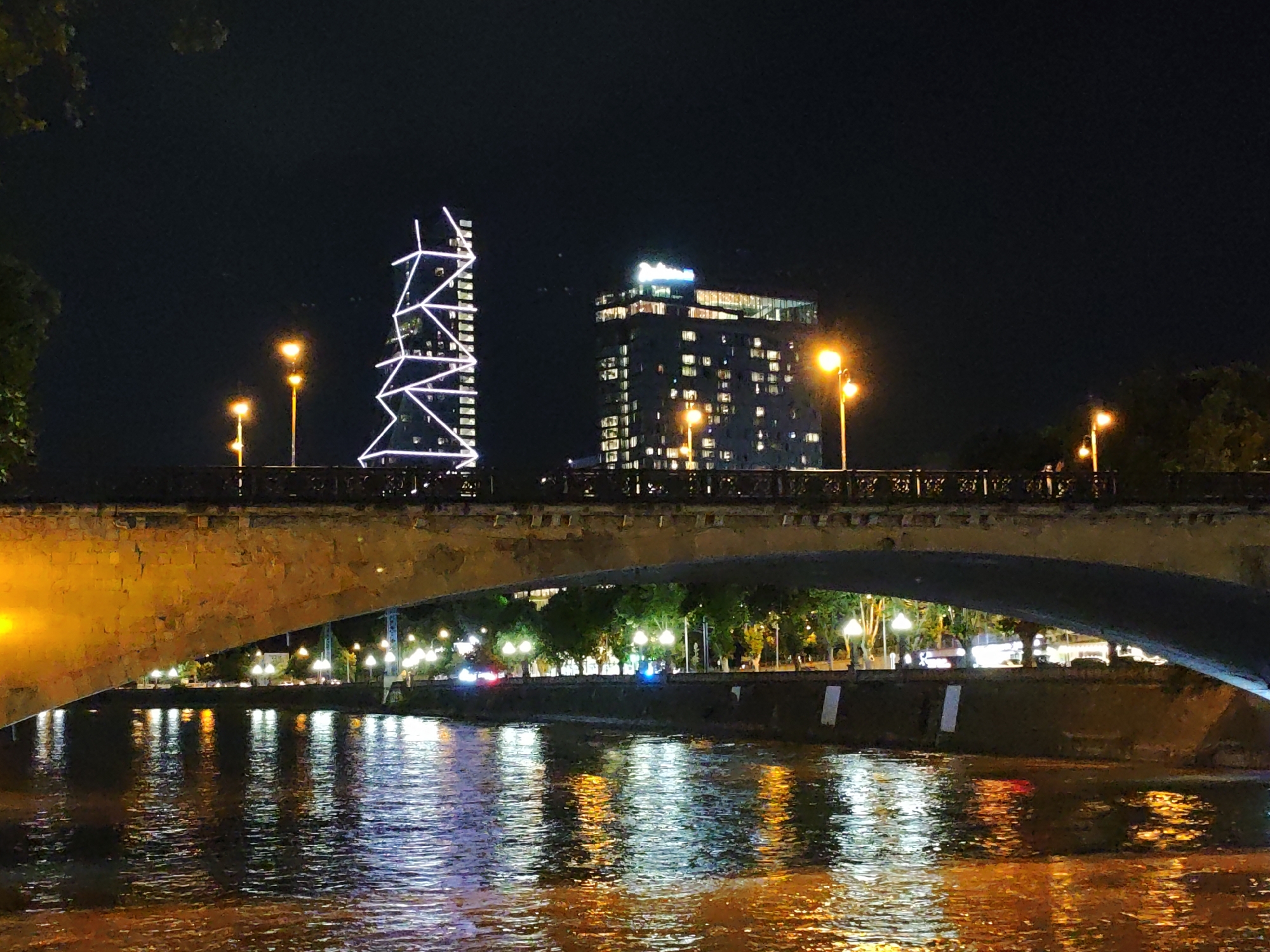 The image captures a nighttime scene in Tbilisi, Georgia, focusing on the Rike Park area, viewed from the Mtkvari River. A large, arched stone bridge dominates the foreground, casting a shadow over the water. The river's surface reflects the lights from the city and the bridge, creating a shimmering effect with warm orange and cool white tones.
Beyond the bridge, two modern buildings stand out against the dark sky. The building on the left is a tall, slender skyscraper adorned with a striking geometric pattern of white lights, resembling a stylized lattice or network. To its right, a wider, multi-story building is illuminated, with numerous windows showing interior lights. The top of this building features a glowing sign that appears to spell out "Radisson."
Several streetlights with orange illumination are visible along the embankment behind the bridge, interspersed with trees that are silhouetted against the cityscape. The overall impression is one of urban nighttime ambiance, with architectural landmarks illuminated and their reflections dancing on the water. There are no visible people or specific actions depicted.