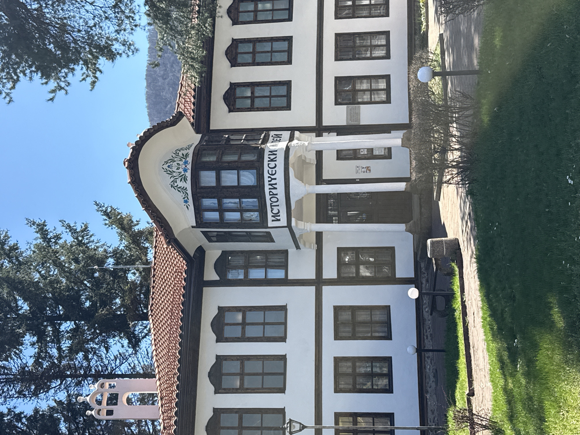 The image depicts the Historical Museum of Etropole, Bulgaria. The building is a two-story structure with a distinctive architectural style, featuring white walls, dark brown wooden accents, and a tiled roof. A prominent bay window adorned with floral designs and the inscription "ИСТОРИЧЕСКИ МУЗЕЙ" (Historical Museum) projects from the front facade. The entrance is marked by a small portico with white columns. The surrounding environment includes trees, grass, and a paved walkway. The day appears to be sunny, with a clear blue sky visible in the background.