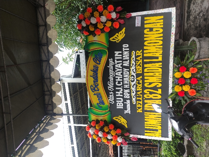 A large, elaborate funeral wreath (papan bunga duka cita) stands prominently, conveying condolences. The wreath features a black rectangular board adorned with vibrant yellow text and colorful floral arrangements at the top and bottom.
The text on the board, which is yellow against the black background, reads: "Turut Berduka Cita" (Deepest Condolences) at the top, followed by "Atas Meninggalnya" (For the passing of). The deceased is identified as "IBU HJ.CHAYATIN," who is further specified as "Ibunda BPK H.HANDY ALIANTO" (Mother of Mr. H. Handy Alianto). The senders of the condolence message are "KELUARGA BESAR" (Extended Family) and "ALUMNI '82 SMADA LAMONGAN," indicating a group of alumni from a high school in Lamongan from the graduating class of 1982. Decorative elements include yellow butterfly outlines and intricate patterns.
The floral arrangements feature dense clusters of red, orange, yellow, and white flowers, with hints of pink and purple blooms, creating a solemn yet colorful display.
The wreath is situated outdoors, possibly under a covered structure with a wavy-edged roof, visible in the background along with green foliage and a clear sky. A black motor scooter is parked in the foreground to the right, partially obscuring the bottom right portion of the wreath. A potted plant, possibly a cactus, can be seen behind the wreath in one of the crop images. The setting suggests a memorial event in Lamongan, Indonesia, during daytime.