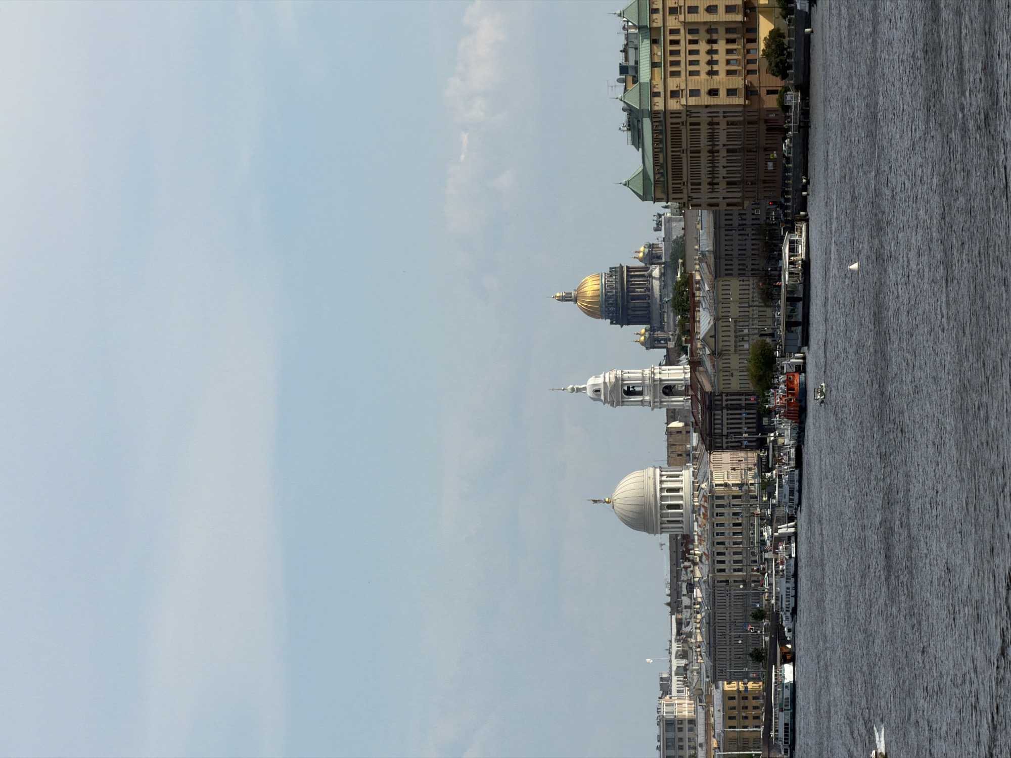 This image presents a panoramic view of the cityscape of Saint Petersburg, Russia, observed across a wide body of water, identified as the Neva River. The sky above is a light blue with scattered, diffuse white clouds covering approximately 40% of the upper frame.
In the foreground, the Neva River occupies the lower half of the image. The water is dark grey-blue, exhibiting a choppy texture with distinct ripples and small waves. A faint white wake is visible in the far left foreground. A small, white, triangular buoy floats in the mid-right section of the river. Further in the distance on the right, a small white motorboat or sailboat is faintly visible, moving across the water. Another small, indistinct object or craft is visible in the far left foreground.
The midground features the densely built urban landscape of Saint Petersburg along the riverbank. Various multi-story buildings, predominantly in shades of pale yellow, ochre, grey, and light brown, line the waterfront. Some buildings display classical architectural elements, while others appear more residential or commercial. Several structures have distinctive green-colored roofs, notably a large building complex on the far right of the cityscape.
Two prominent cathedrals dominate the skyline. In the mid-right of the city, St. Isaac's Cathedral is clearly identifiable by its large, central golden dome and several smaller golden domes. To the left of St. Isaac's, approximately centered in the midground of the city, stands the St. Nicholas Naval Cathedral (Nikolsky Cathedral), characterized by a prominent white classical dome with columns and a tall, slender, multi-tiered white bell tower positioned immediately to its right.
Along the river embankment, multiple docks and piers are visible. Several large, white passenger boats or river cruise vessels are moored along these docks. A single bright red boat is also docked further to the right. Small figures, likely people, are visible on the docked boats and along the embankments, but they are too distant for detailed identification. The overall scene depicts a serene daytime view of a historical European city.