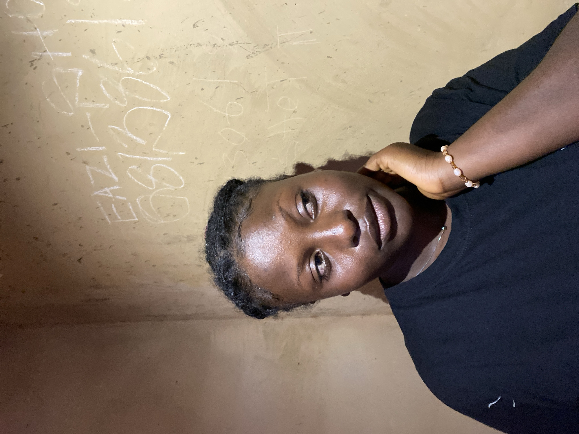 A medium, close-up shot depicts a dark-skinned woman indoors, presumably in Kuru Dungus, Nigeria. She has dark, braided hair with edges, wears a black t-shirt, and a beaded bracelet. Her right hand rests on the side of her neck. The background features a beige-colored wall with some faded, illegible writing on it. The lighting is dim and uneven.