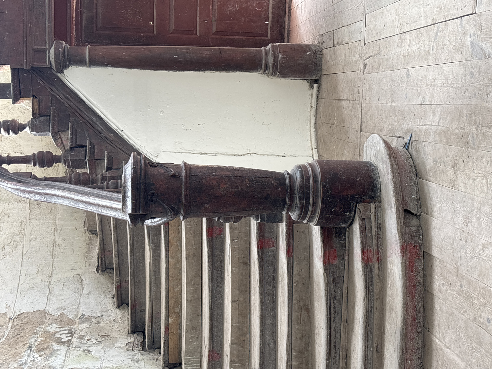 The image depicts a weathered, wooden staircase and surrounding interior elements in a dimly lit, possibly abandoned or historical building. The staircase is curved, with prominent dark wood banisters and balusters, some of which show signs of age and wear. The steps themselves appear to be constructed from a lighter material, possibly concrete or stone, with visible red markings on the front edges of some. The walls flanking the staircase are textured and discolored, with peeling plaster. To the right, a section of a dark red wooden door is visible, bearing what appears to be faint, handwritten inscriptions. The floor in front of the staircase is made of light-colored wooden planks, also showing signs of age and wear. The overall impression is one of a neglected or historic space. This scene is located in Жалудоцкі сельскі Савет, Belarus.
