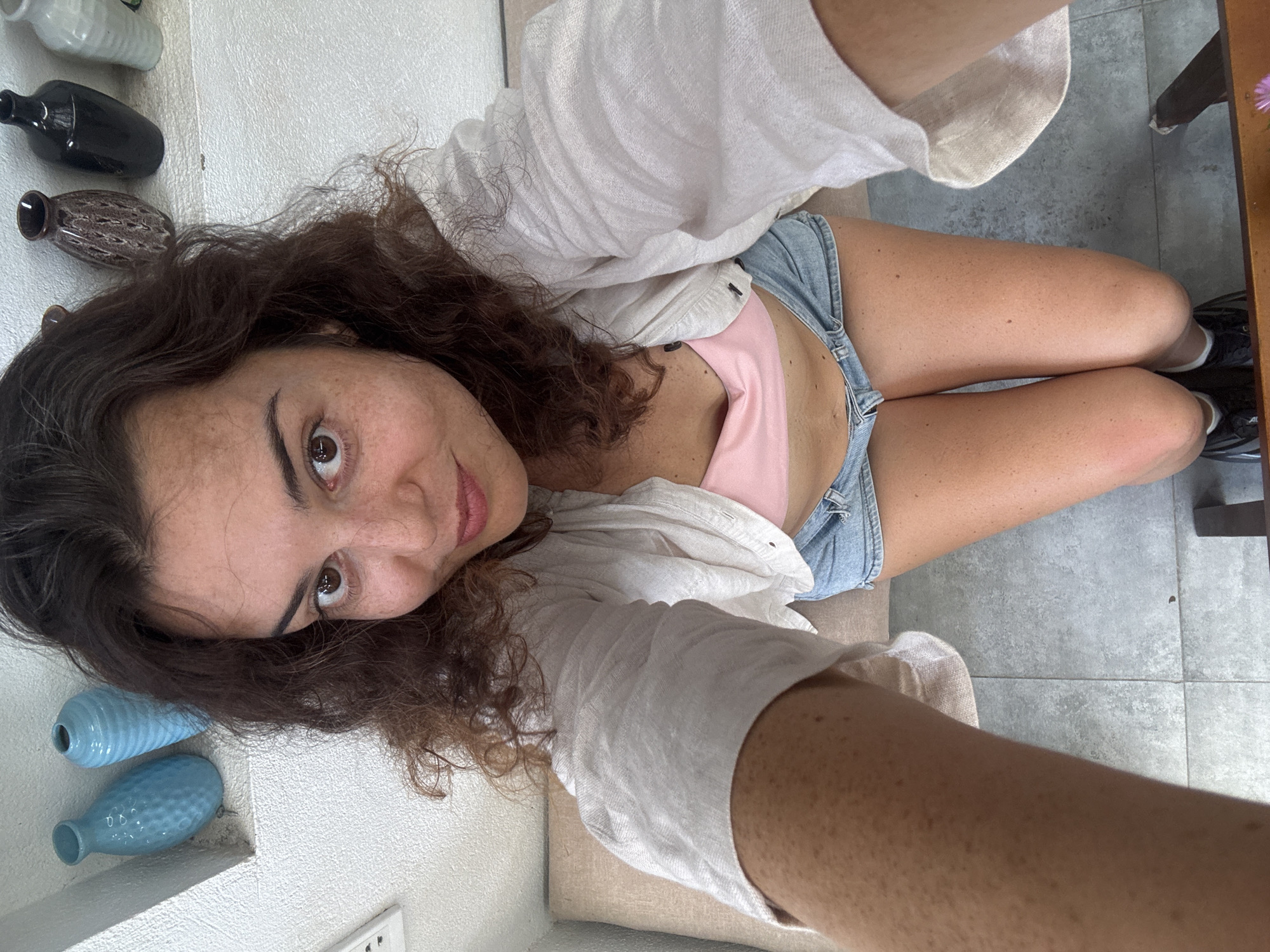 A young woman with curly brown hair and freckles is kneeling on a tiled floor in what appears to be an indoor setting. She is wearing a pink bikini top and denim shorts, with a light-colored, open-buttoned linen shirt over the top. The background shows a white textured wall with a shelf holding several decorative vases. Two blue vases are on the left side of the shelf, and a brown, black, and white vase are on the right. The woman is looking directly at the camera, with her right arm extended. The scene suggests a casual, possibly residential or boutique environment. The specific location in Hoi An, Vietnam, is not discernible from the visual information provided.