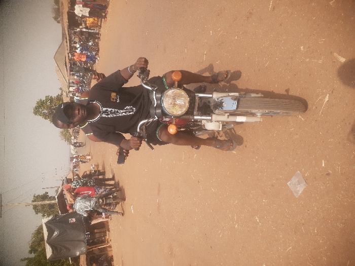 An outdoor scene in Gwadanye, Nigeria, depicts a bustling street or market environment under clear daylight. A dark-skinned male operates a red and chrome motorcycle in the foreground, looking directly at the viewer. He wears a black short-sleeved top with white embroidery, black shorts, a black baseball cap with a white logo, and dark sandals. Behind him, a partially visible dark-skinned male passenger wears a striped tank top and a black cap. The motorcycle's front features a circular headlight and orange indicator lights. The road surface is unpaved, composed of reddish-brown earth with lighter patches and scattered debris; a clear plastic wrapper is visible on the ground to the left of the motorcycle's front wheel. The midground on both sides of the road displays makeshift stalls, some covered with black tarps, and permanent structures with corrugated metal roofs. Numerous dark-skinned individuals, dressed in various casual clothing, are present, some standing near motorcycles or gathered in groups. Additional motorcycles are visible throughout the midground. The background shows distant buildings and sparse trees against a light, hazy sky, with utility poles on the left.