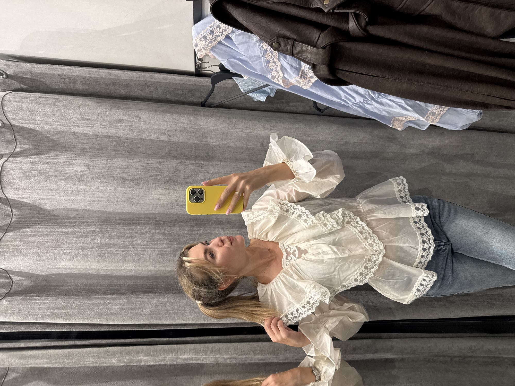 A woman with blonde hair pulled into a ponytail takes a selfie in a room with a reflective surface, possibly a mirror or highly polished floor, at the bottom of the frame. She is wearing a white blouse with ruffled sleeves and lace trim, and blue jeans. She holds a yellow phone in her right hand. Behind her, several garments are hanging on hangers: a brown leather jacket and a light blue dress with lace trim. The background consists of a textured, grey fabric. The lighting suggests it is daytime.