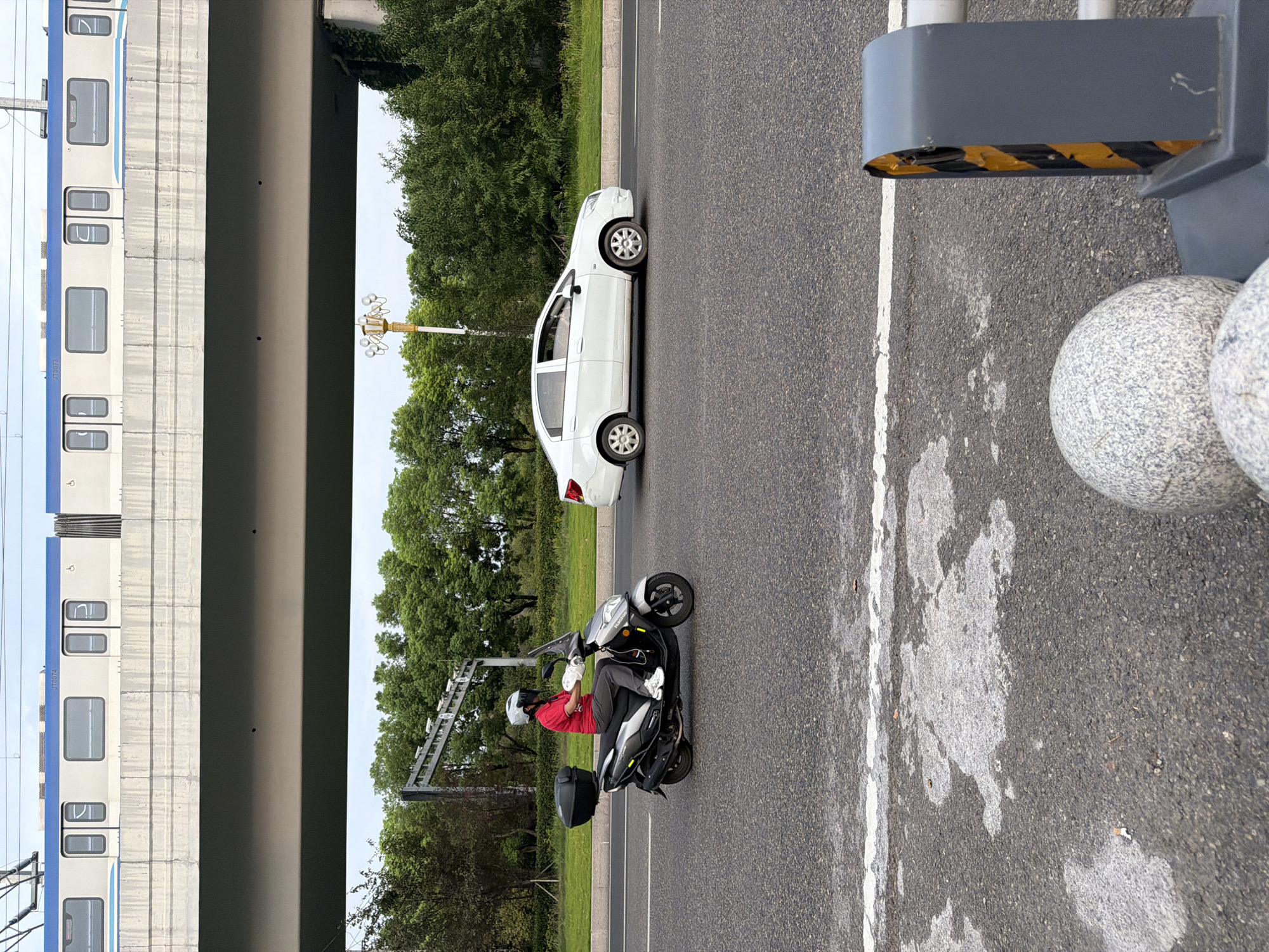 In the city of Jiangjin, China, a person wearing a red shirt, white helmet, white gloves, and white shoes is riding a silver scooter with a black storage box on the back. The scooter is in the left lane of a two-lane road. A white sedan is in the right lane, ahead of the scooter. In the background, there is a train elevated on a concrete structure, with trees and greenery beyond the road. The sky is overcast. The road surface is asphalt with white lane markings. There are some white patches on the road that appear to be water or residue. In the foreground, there are two spherical stone bollards and a gray traffic barrier.