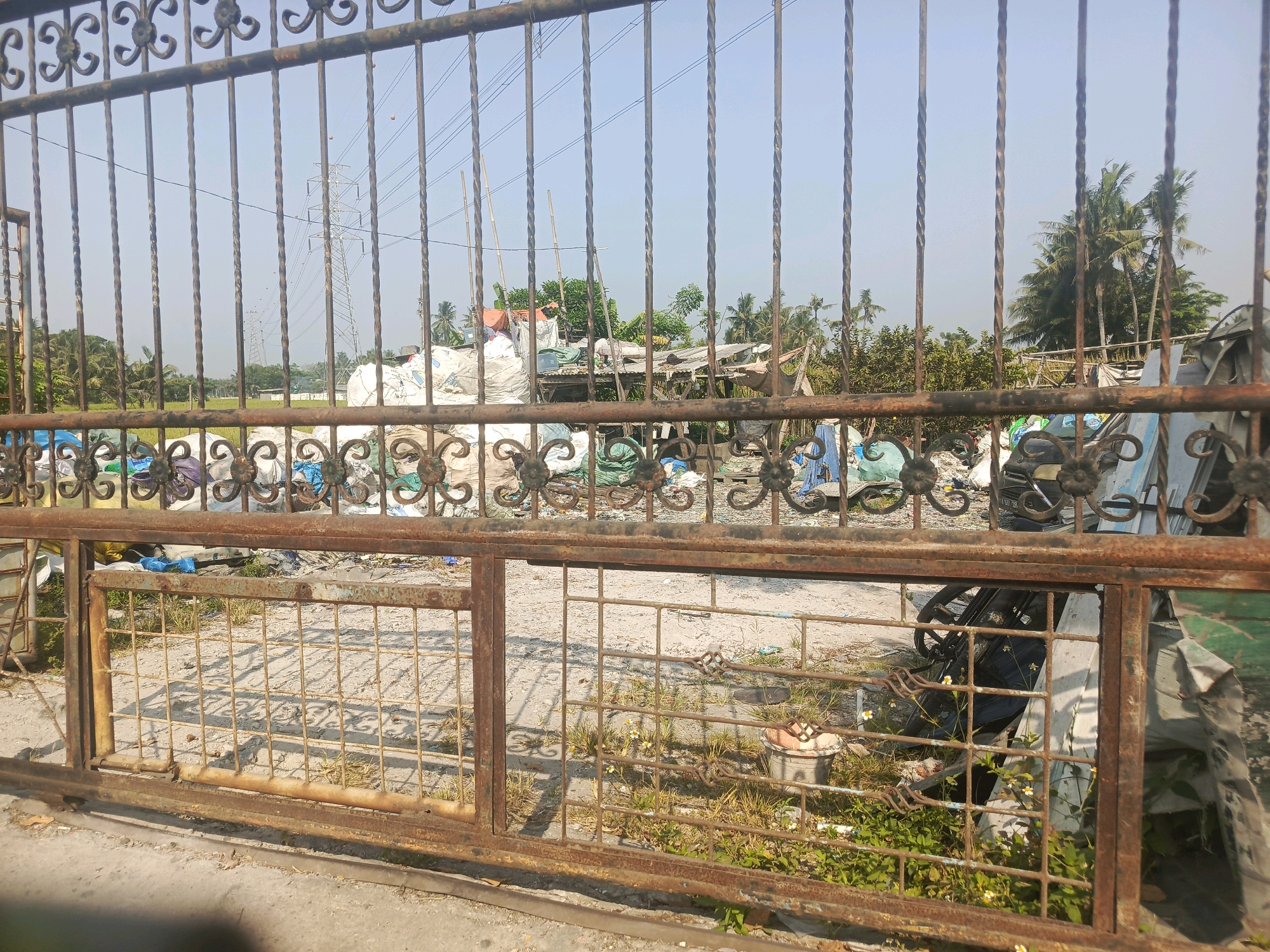 A rusty metal gate in Rawa Boni, Indonesia, with a cluttered area behind it. The area behind the gate appears to be a dumping ground, filled with large white sacks, blue tarps, and other debris. In the background, there are palm trees and power lines. The sky is clear and blue. There are no people visible in the image.