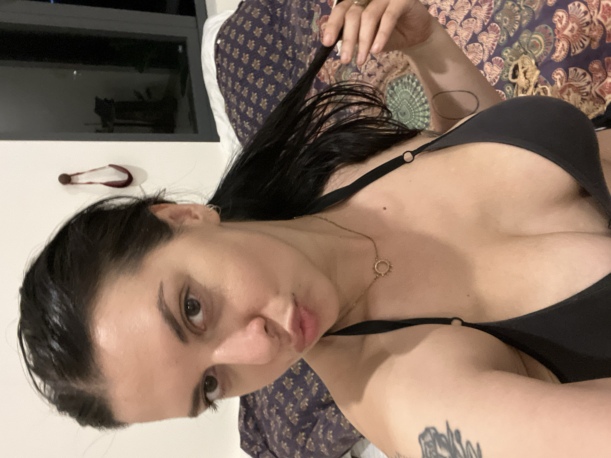 The media file shows a close-up, tilted shot of a person in a bedroom setting. The person has long, dark hair and is wearing a black bikini top. They are lying down on a bed with a patterned duvet. A small, circular necklace with a sun-like charm is visible. The person's left arm is extended, with a tattoo visible on their forearm. The background includes a window and a white wall with a small object that appears to be a hair clip. The lighting suggests it is likely daytime. The person's facial expression is neutral. The location is identified as Đà Nẵng, Vietnam.