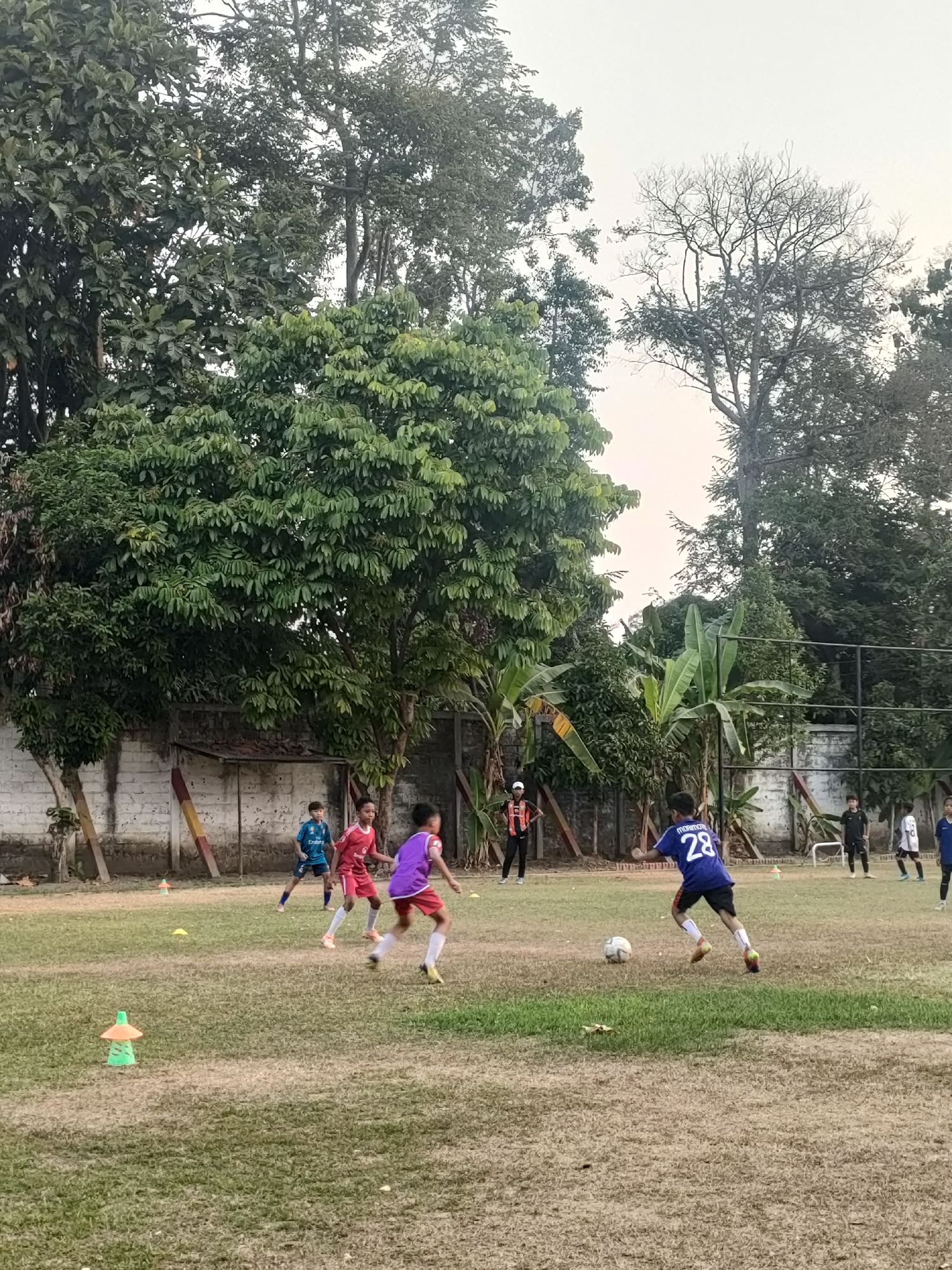 The image depicts a group of young boys playing soccer on a dry grass field in Banguntapan, Indonesia. The field is surrounded by lush green trees and a low concrete wall. In the foreground, a boy wearing a blue jersey with the number 28 on the back is dribbling a soccer ball. Other children in red and purple jerseys are also visible on the field, engaged in the game. In the background, a few adults are standing, possibly coaching or observing. The sky is overcast, suggesting a cloudy day. The scene captures a moment of youthful athleticism and camaraderie in a tropical setting.