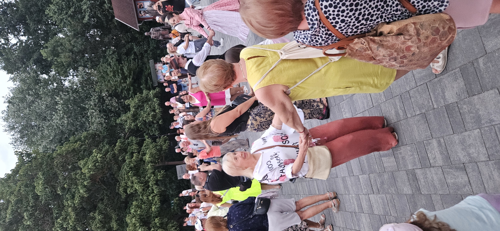 The image depicts an outdoor gathering of people, likely a public event or festival, taking place on a paved area in the city of Hévíz, Hungary. In the foreground, two women are dancing in a circle, holding hands. The woman on the left wears a white shirt with a graphic print and red pants, and carries a light-colored crossbody bag. The woman on the right is dressed in a bright yellow sleeveless dress and has a cream-colored backpack slung over her shoulders. Behind them, a larger group of people are also engaged in dancing or observing, forming a semi-circle. Several individuals are captured in motion, some in traditional attire, such as a man in a vest and fedora, and a woman in a pink and white dress. The background is characterized by lush green trees, and a small building with a red roof is visible to the right. The ground surface is made of interlocking grey pavers. The overall atmosphere suggests a festive and communal activity.