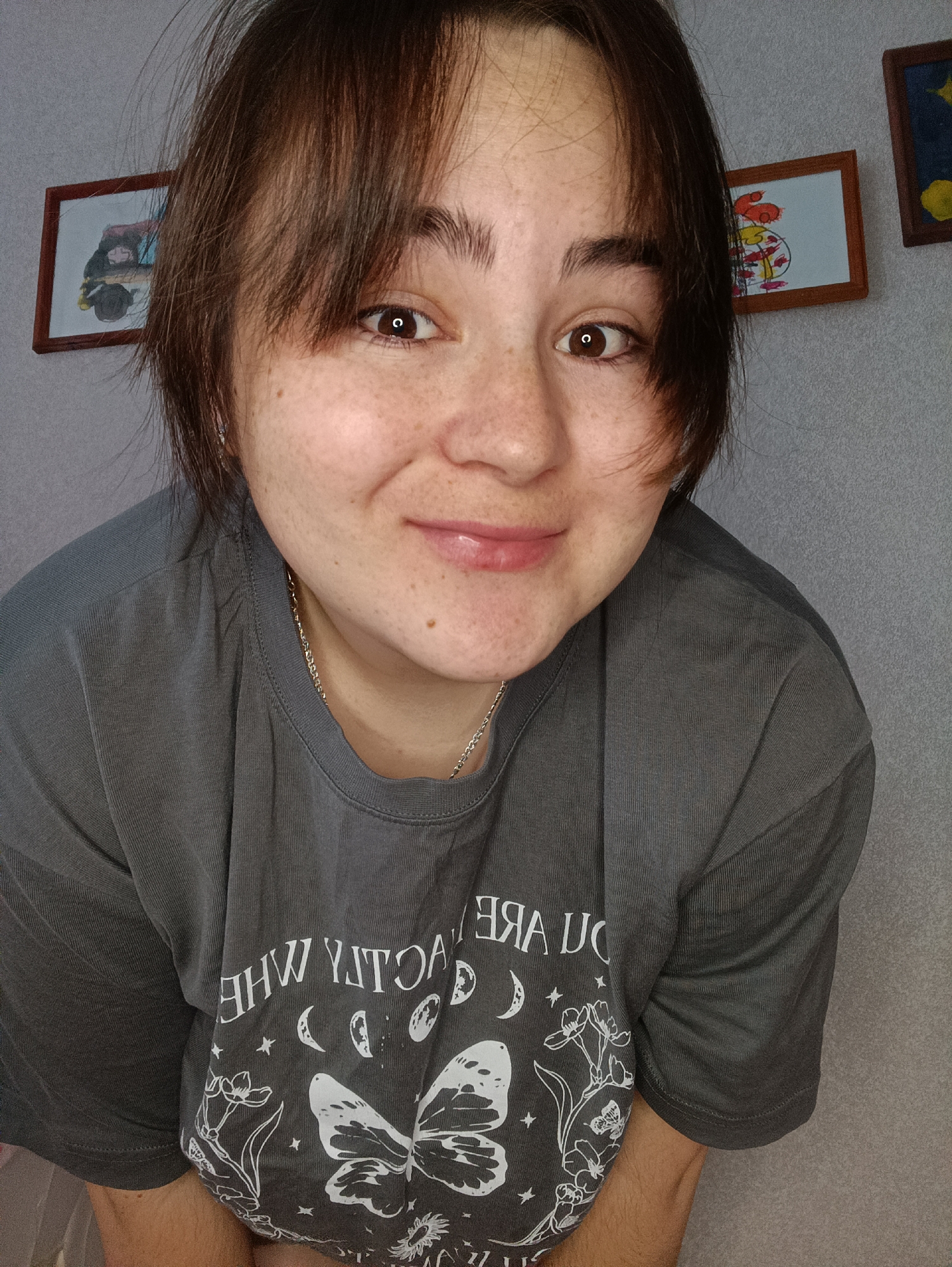 A close-up selfie features a fair-skinned person with brown hair and bangs, freckles, and brown eyes. They wear a gray T-shirt with a white butterfly and celestial design, as well as a silver chain necklace. Behind the person, a gray wall is decorated with three framed pictures. The location of this image, specified as the city of Noviy, Russia, is irrelevant to the visual content.