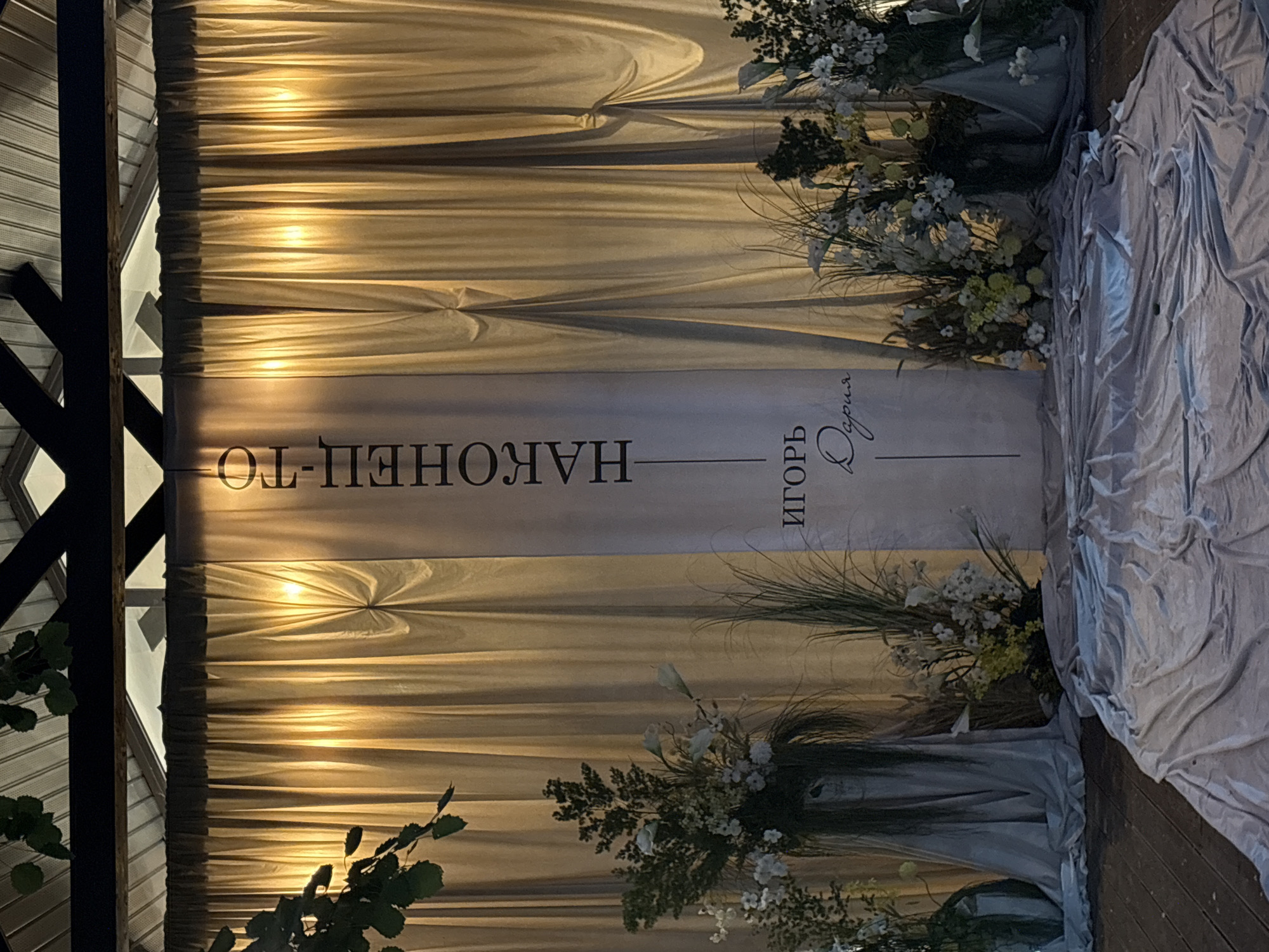 This image captures a decorated event space, likely an aisle or altar setup for a ceremony, under a semi-enclosed structure. A large, vertical white banner serves as the central focal point against a backdrop of luminous golden-beige curtains. The banner displays the Cyrillic text "НАКОНЕЦ-ТО" (Finally) at the top, followed by the names "Игорь" (Igor) in a sans-serif font and "Дария" (Daria) in a cursive script, connected by a thin vertical line.
The golden-beige curtains are evenly pleated and illuminated from behind by multiple light sources, creating distinct bright vertical glows. Above the curtains, a dark-colored metal beam structure supports a light-toned, corrugated roof. Glimpses of an outdoor environment or sky are visible through gaps in the roof structure. Green leaves from an overhead plant partially intrude into the upper left quadrant of the frame.
The floor consists of dark wooden planks, covered in the foreground by a heavily wrinkled, light grey fabric, suggesting a designated path or area. Flanking the central banner are elaborate floral arrangements. The left arrangement features white flowers, including apparent calla lilies and small white blossoms, amidst dense green foliage and long, dark grasses, set in a dark container partially draped with the same light grey fabric found on the floor. The right side showcases similar arrangements with white and light yellow flowers, various green plants, and tall, wispy grasses, also incorporating dark containers and draped grey fabric. No individuals are present in the scene. The image provides no specific visual cues to identify a precise location within the city of Perm, Russia.